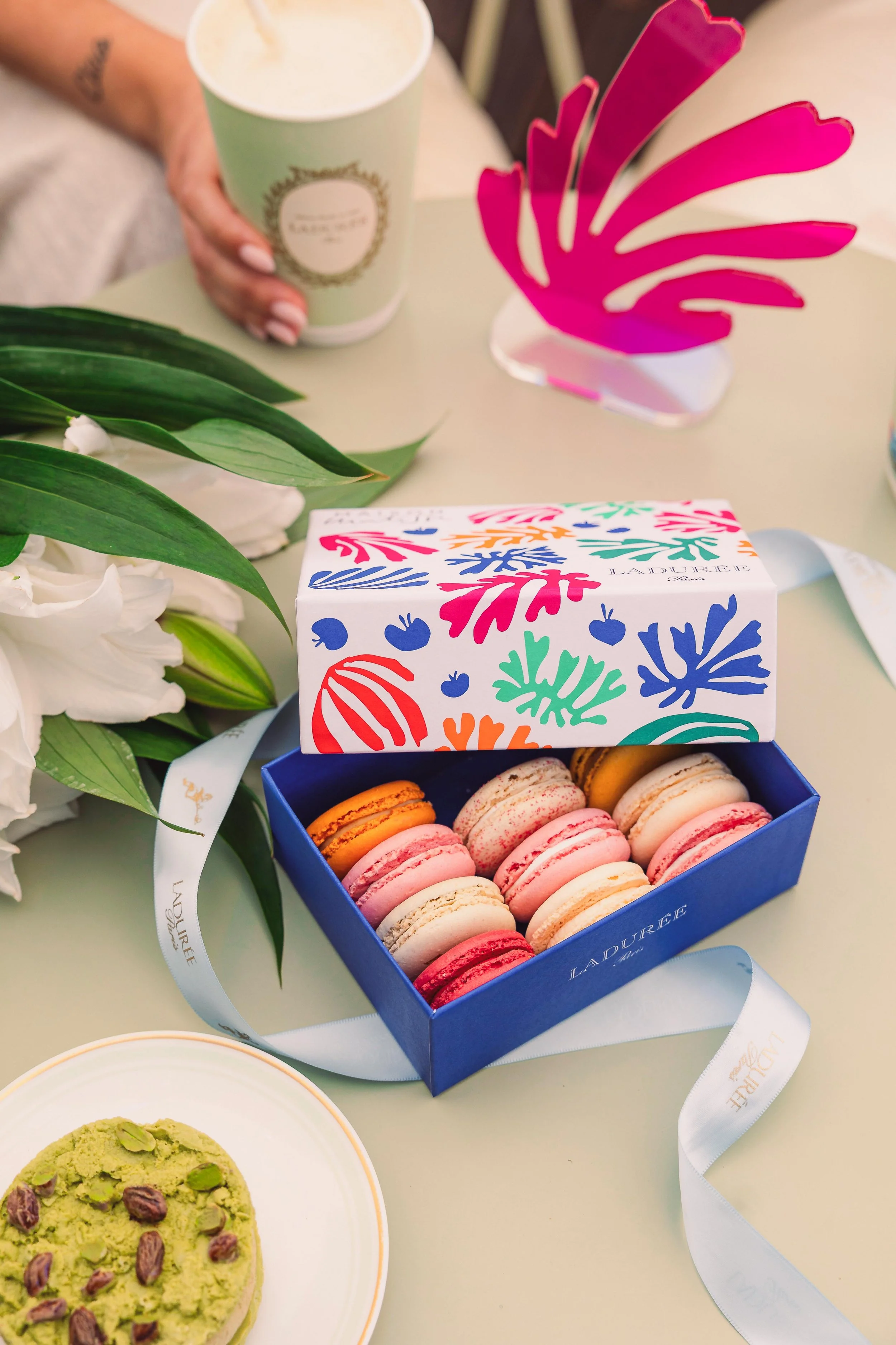 A box of colorful macarons on a table with a cup of coffee, a green plant, flowers, and decorative pink paper in the background.