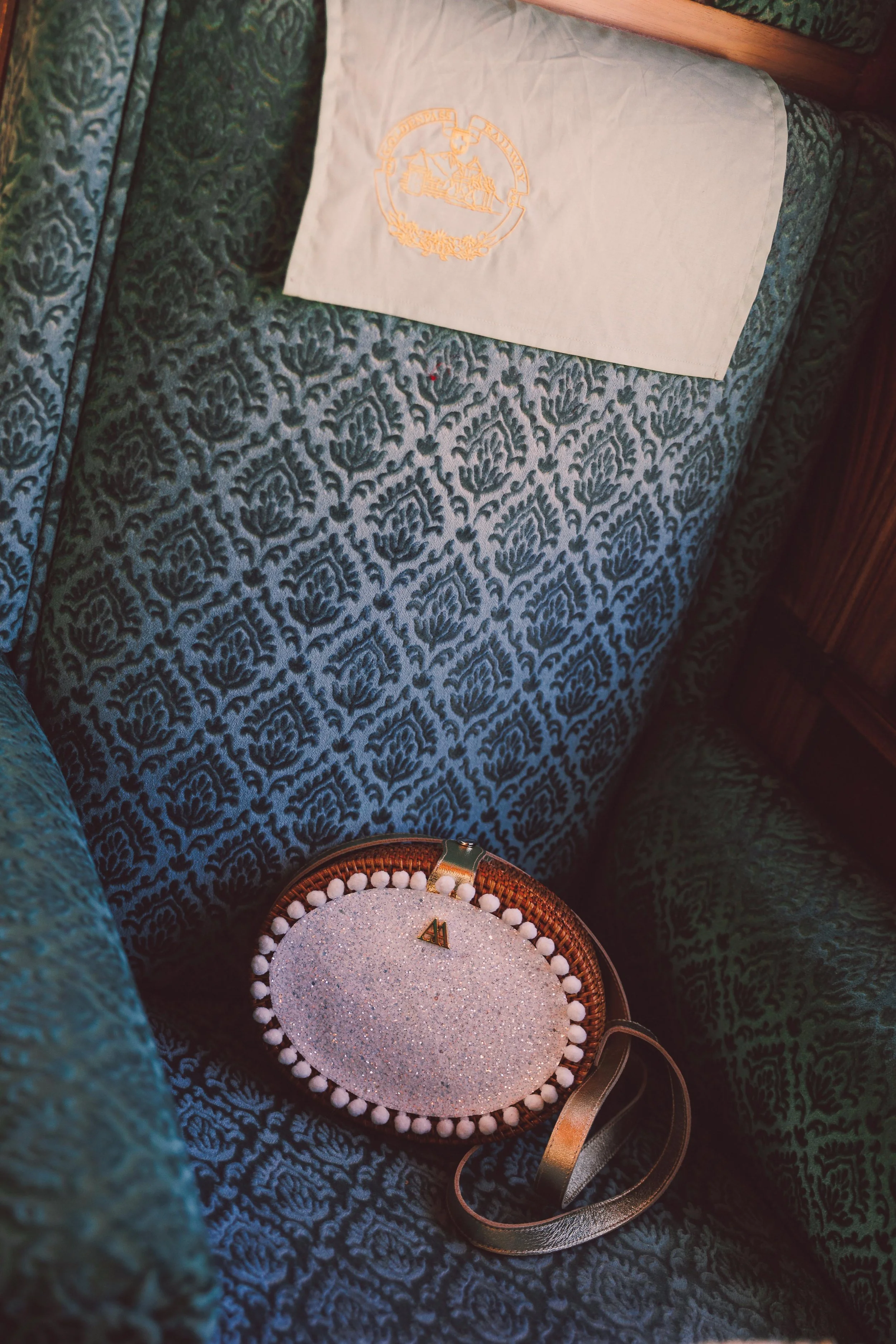 A cushioned green armchair with a white embroidered napkin or cloth resting on its back, a glittery oval clutch purse with pom-pom trim and gold accents placed on the seat, and a metallic strap extending from the purse, with part of a wooden side tab
