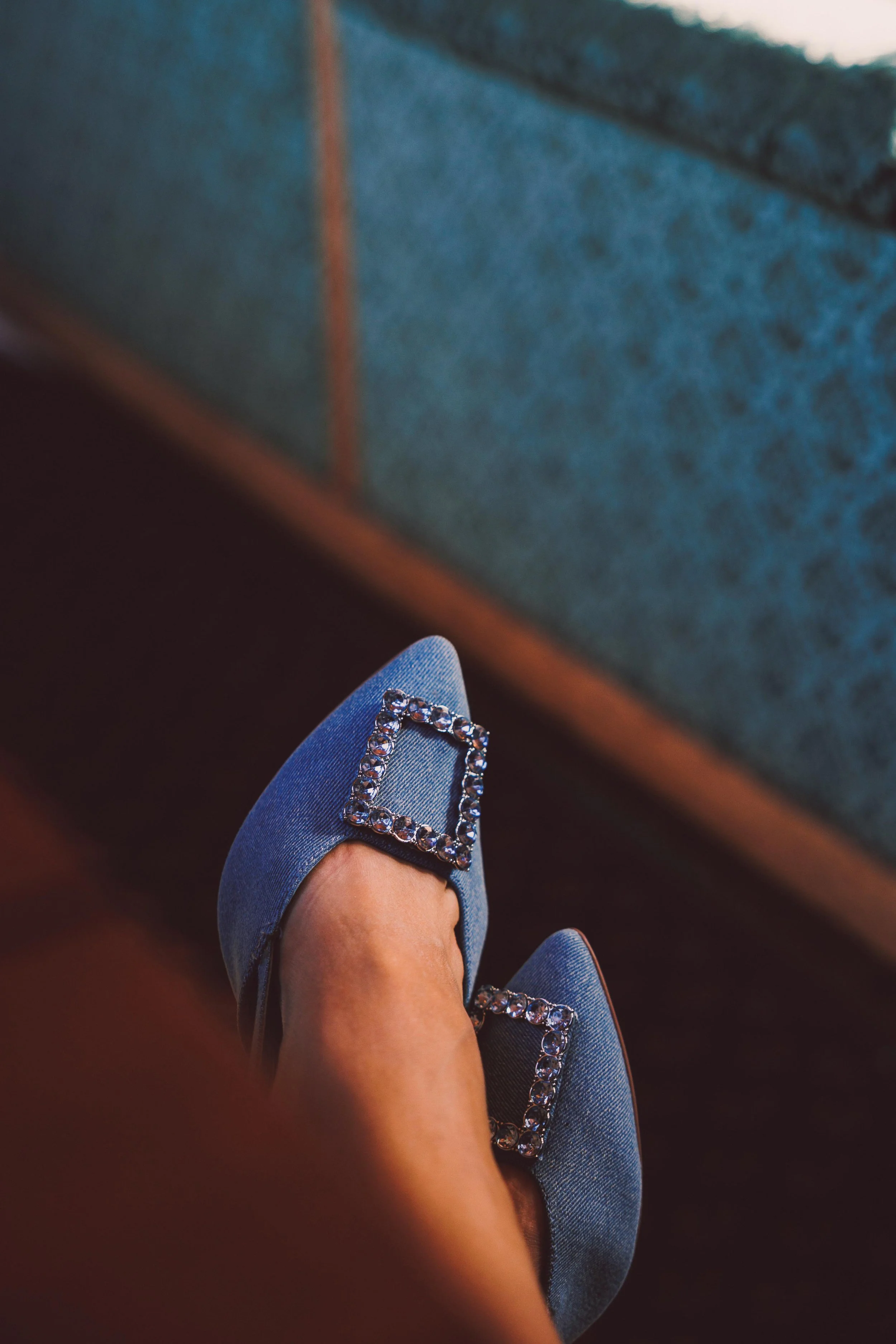 Close-up of a person's legs and feet wearing blue high heels with large square rhinestone embellishments, sitting on a wooden surface near a blue upholstery piece.