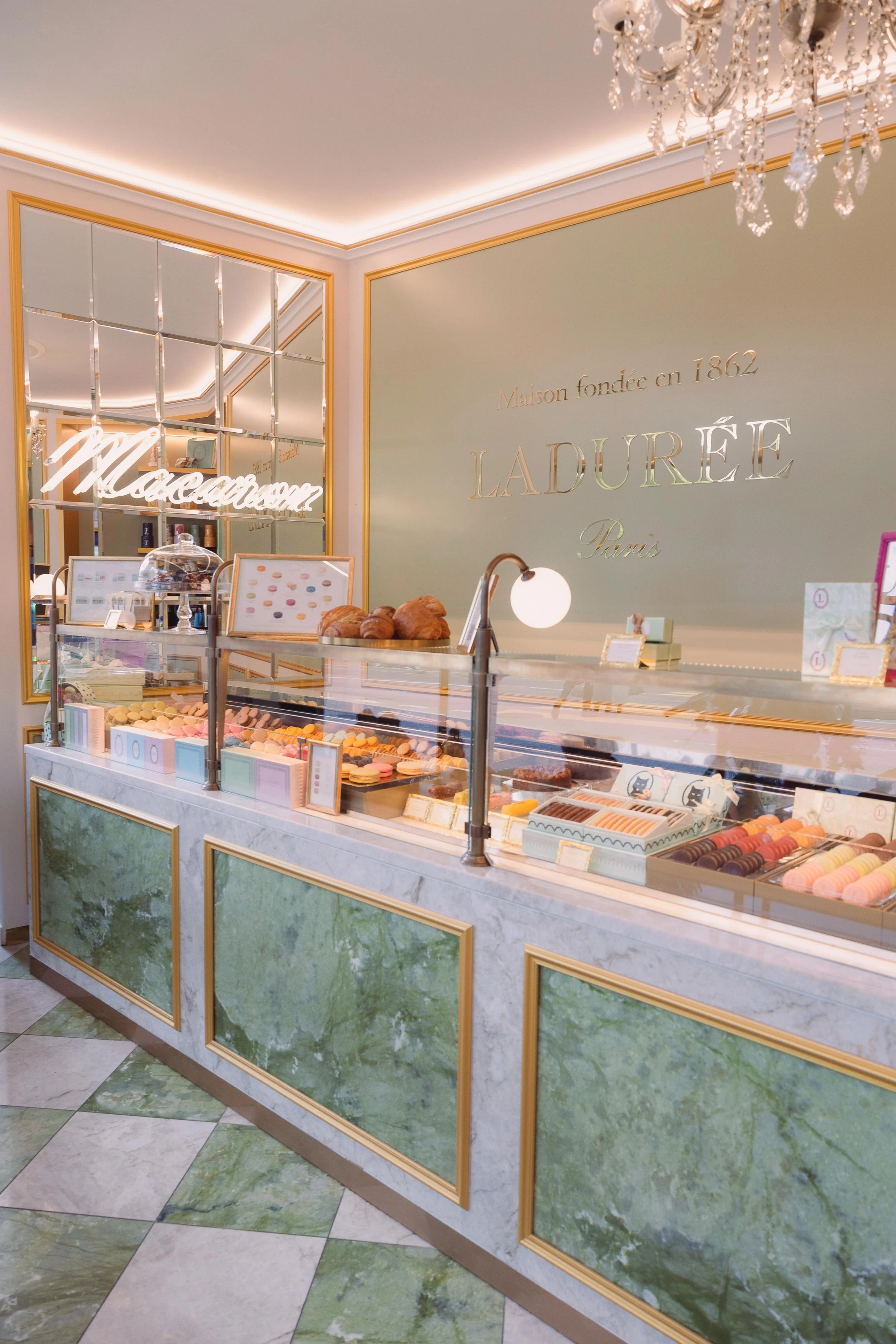 Interior view of a luxurious cake and pastry shop named Ladurée, featuring a marble counter displaying colorful macarons and croissants, with a chandelier hanging overhead and a mirror reflecting the shop's branding on a pastel green wall.