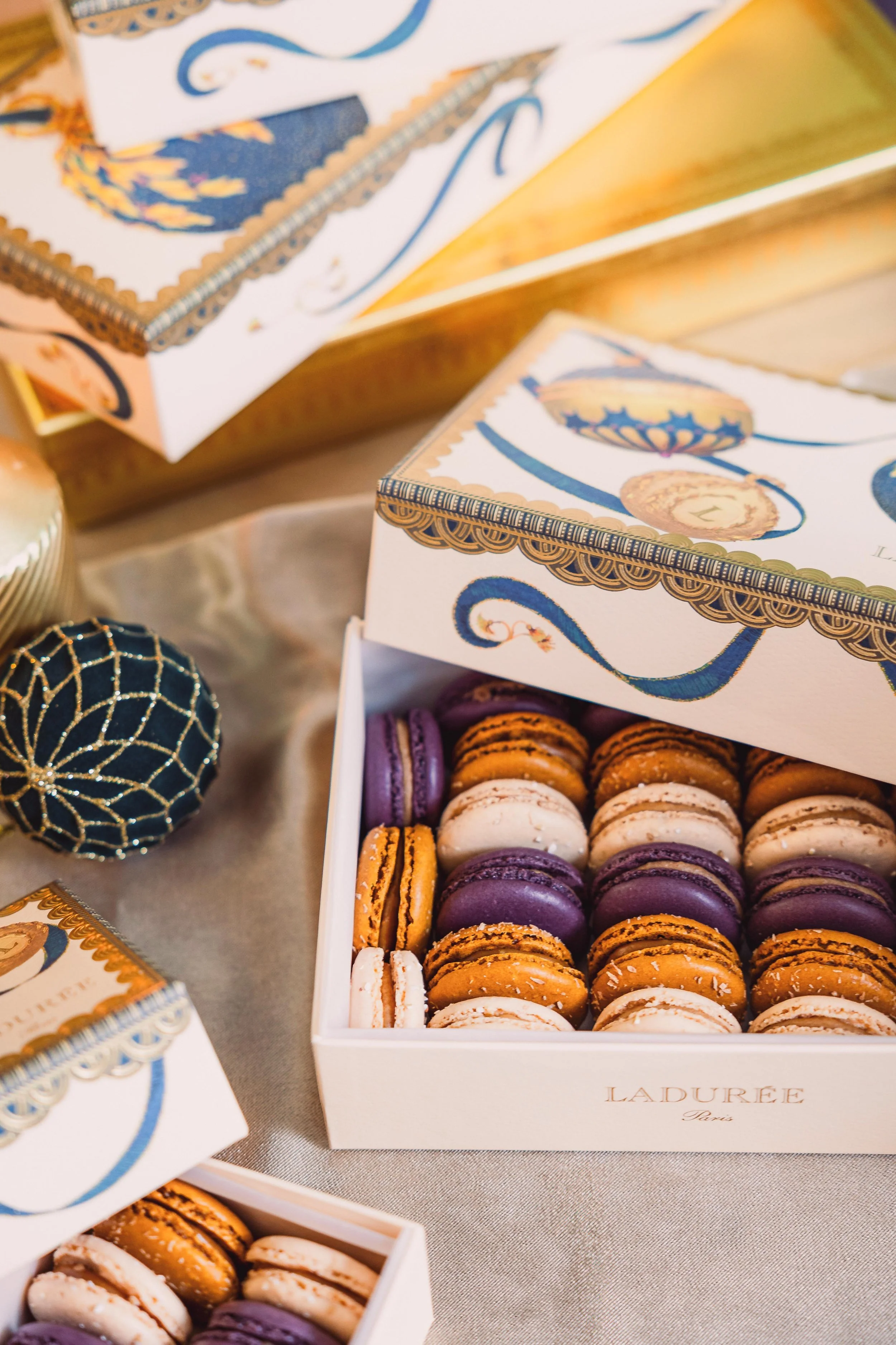 Box of colorful macarons in purple, orange, and white inside a decorative Laduree Paris box, with Christmas ornaments nearby, on a table.