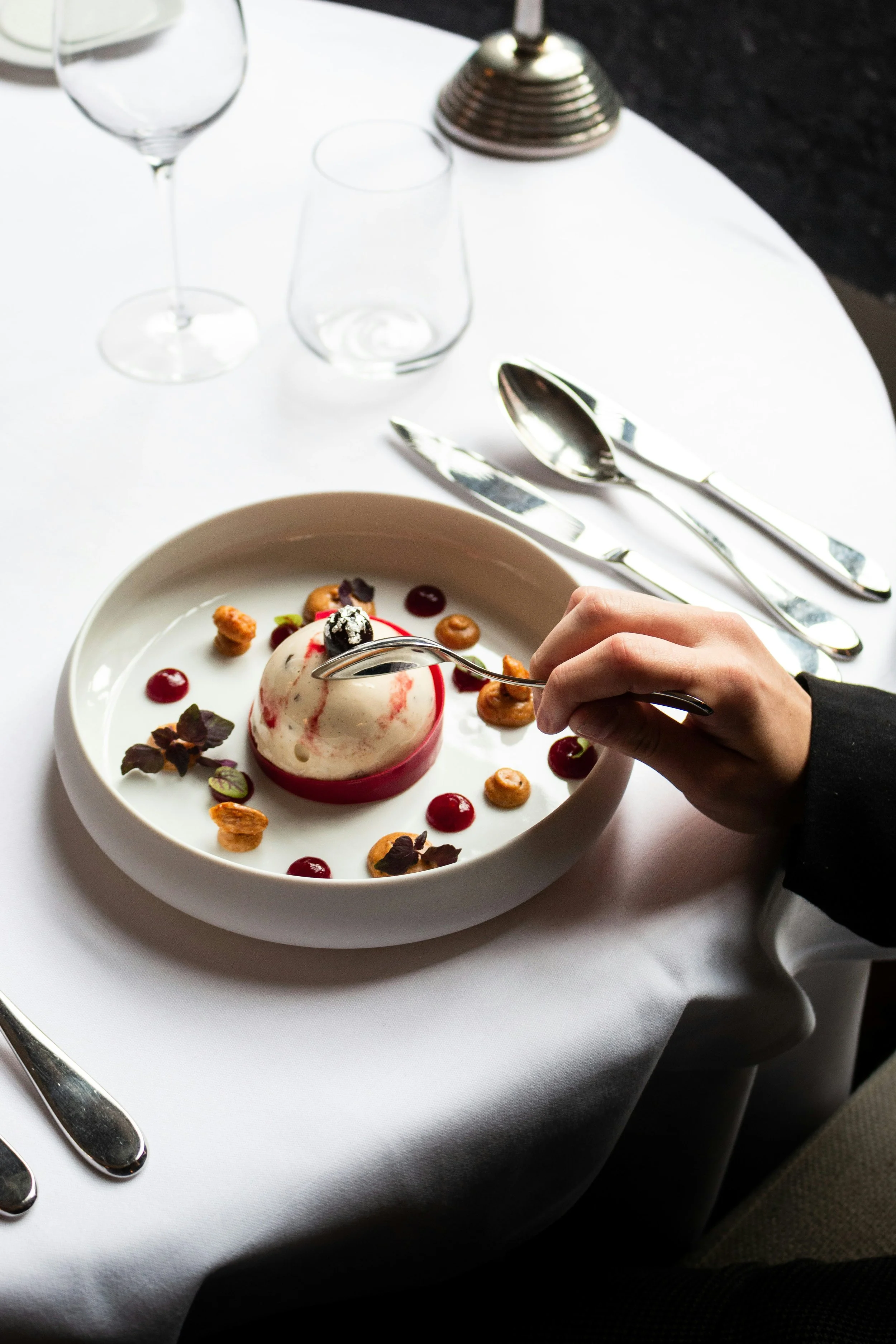 A person holding a spoon over a plated dessert with a white and red spherical shape, garnished with small edible flowers and dollops of sauce, on a white tablecloth with silverware and empty glasses nearby.