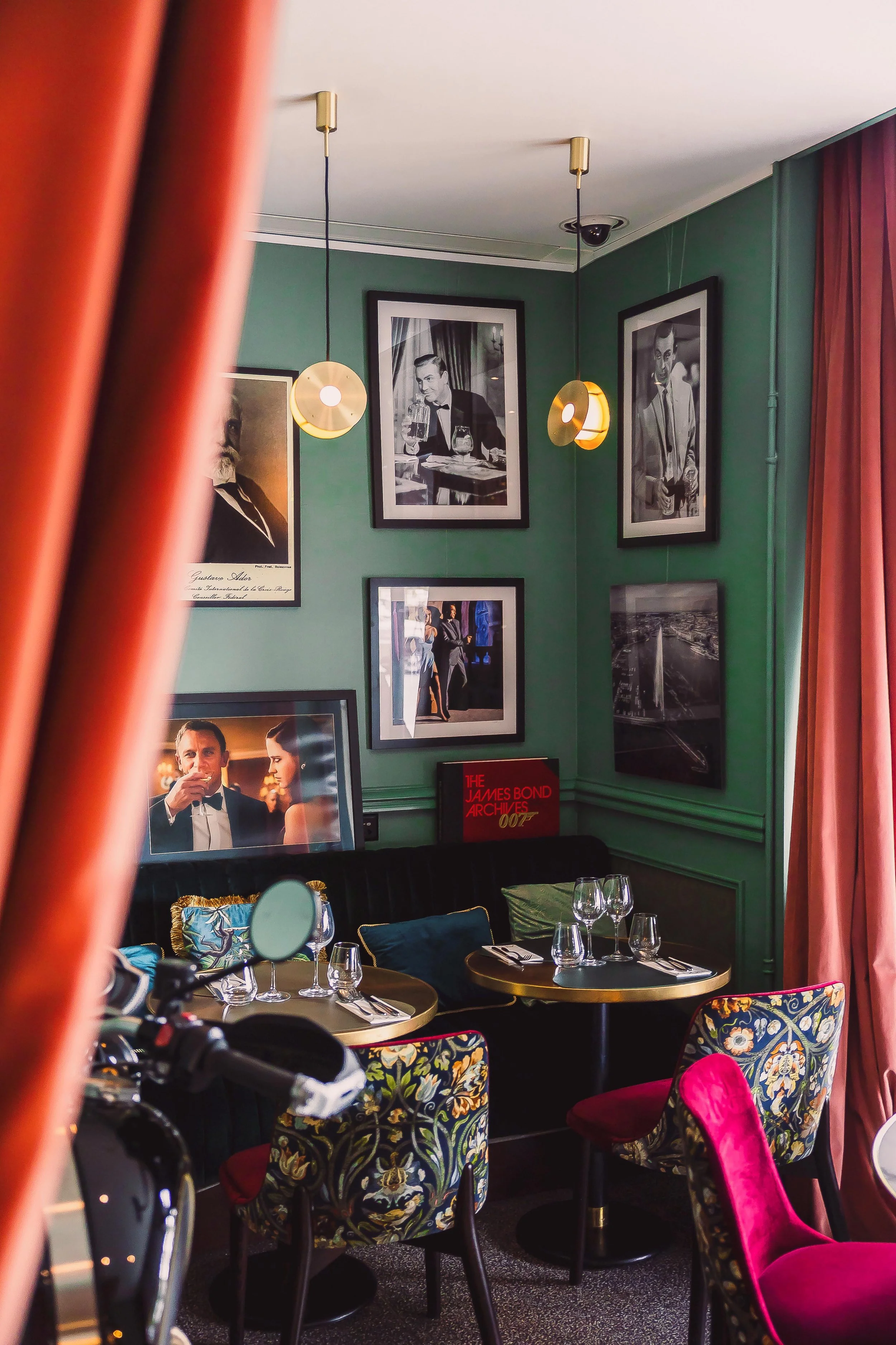 A cozy corner of a restaurant with green walls, black and white framed photos of James Bond actors, a colorful floral-patterned chair, a small table set with glassware and cutlery, pink curtains, and a flat-screen TV showing a scene from a James Bond