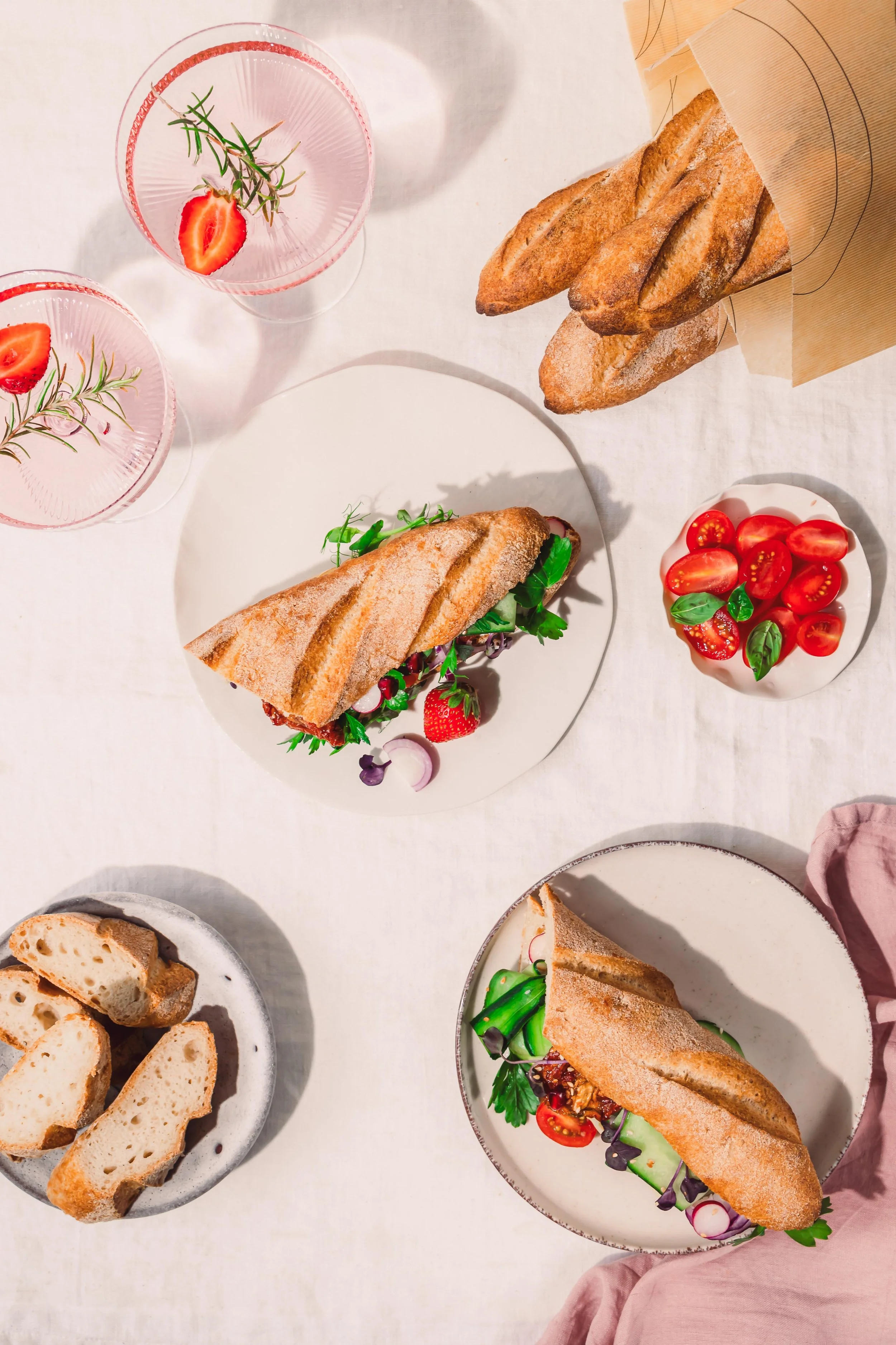 A table with two sandwiches, a small bowl of cherry tomatoes and basil, sliced bread, and two pink drinks garnished with strawberries and rosemary.