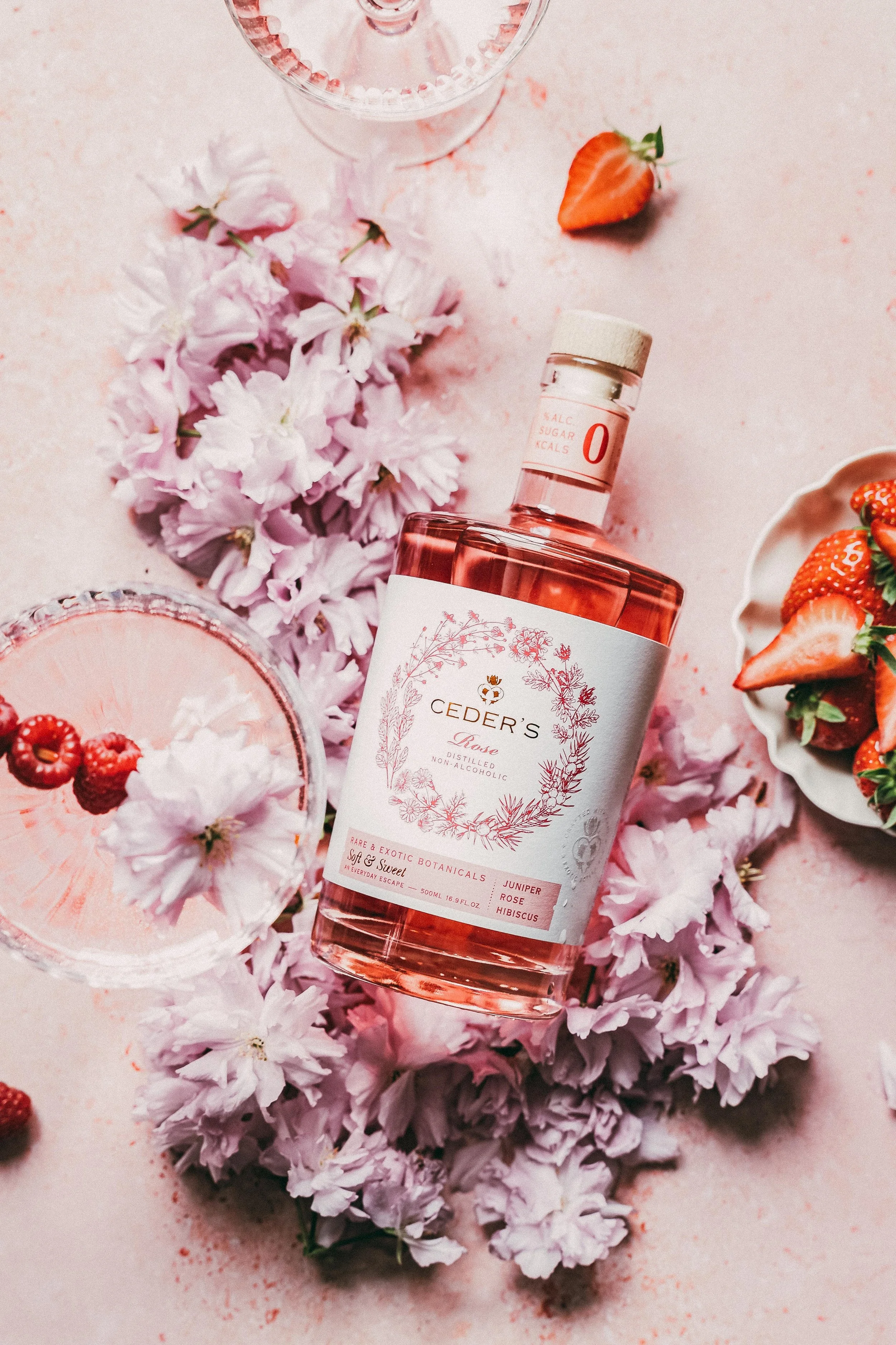 Bottle of Ceder's Ros, non-alcoholic rose-flavored spirit surrounded by pink flowers, strawberries, and a glass of pink drink with berries, on a pink surface.