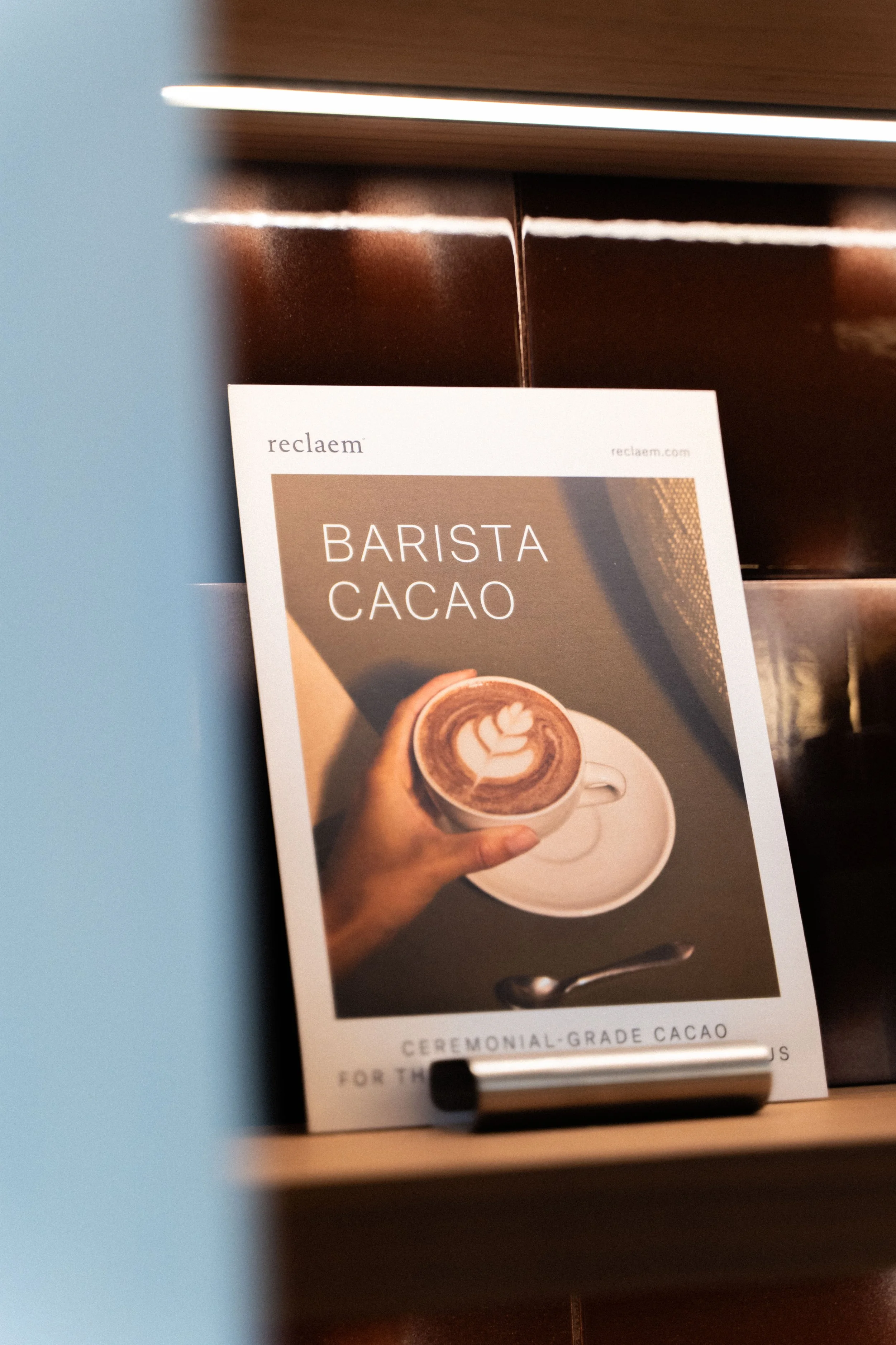 A menu or flyer titled 'Barista Cacao' showing a hand holding a cup of hot chocolate with latte art, on a saucer with a spoon.