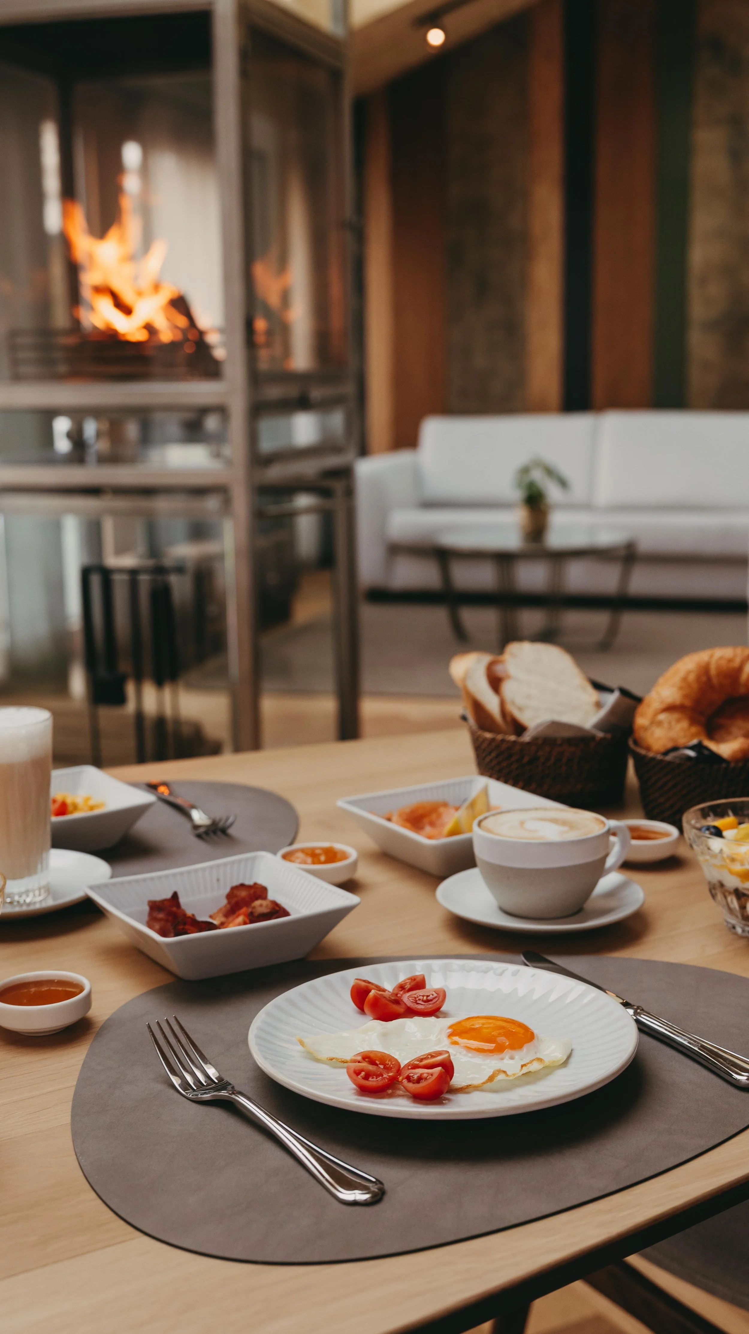 A breakfast table with a sunny side up egg topped with cherry tomatoes, several small dishes of sauces, crispy bacon, pastries including croissants and bread, a cup of coffee with foam art, and a glass of latte, set in a cozy dining area with a wood 