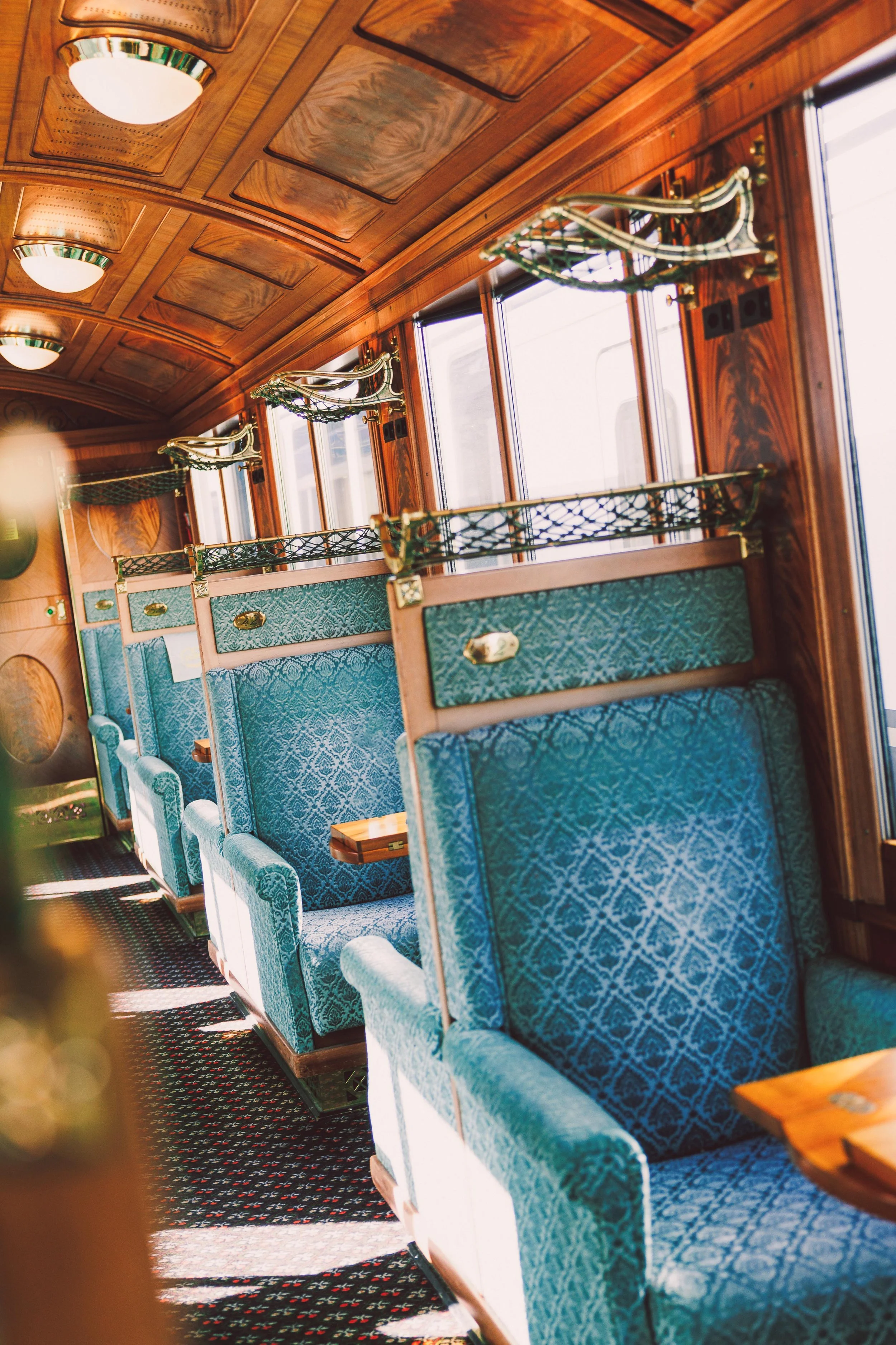 Inside of a vintage train car with teal patterned seats, wooden paneling, windows, and overhead luggage racks.