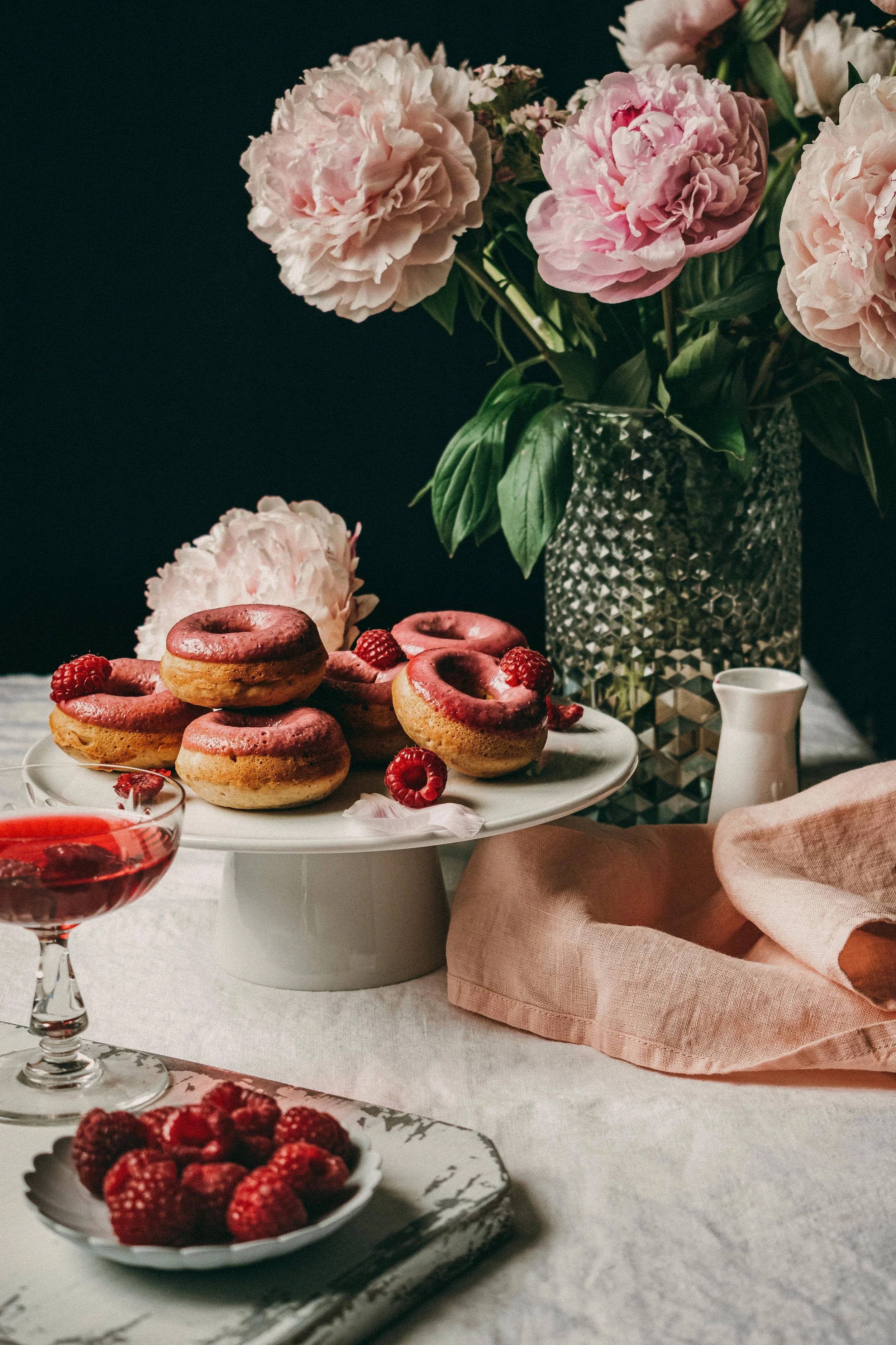 A table with a bouquet of pink peonies in a textured gray vase, a white cake stand with donuts topped with pink icing and raspberries, a glass of red beverage with raspberries, a small white plate of raspberries, a pink napkin, and a white ceramic pitcher against a dark background.