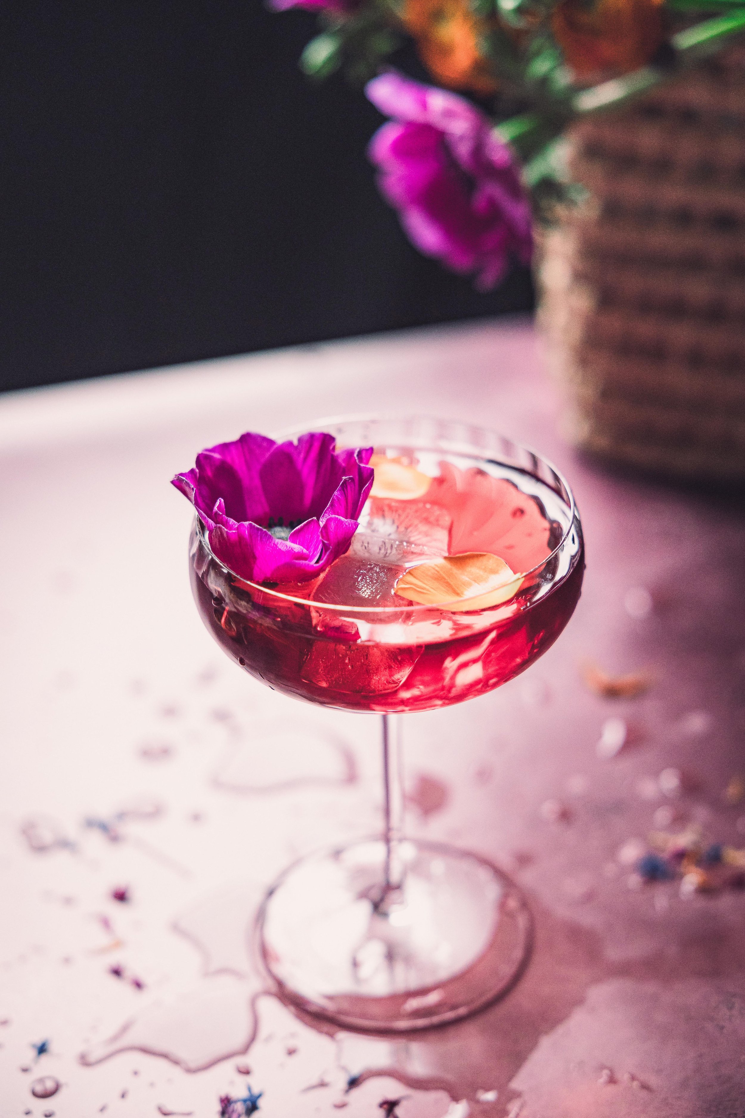 A pink cocktail in a coupe glass with edible flower petals and a purple flower garnish, on a pink surface with a blurred background of a wicker basket with flowers.
