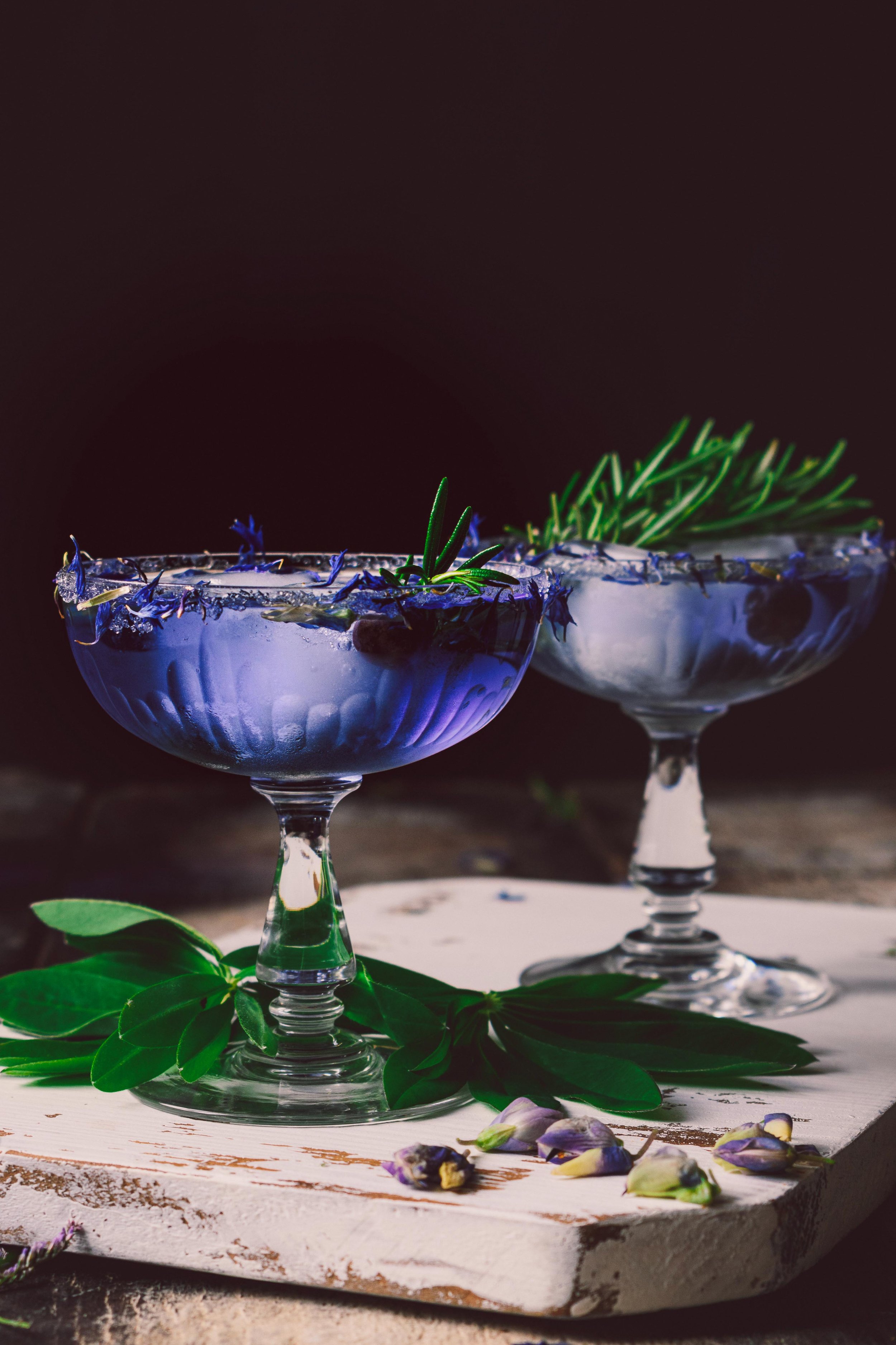 Two glass bowls with blue and white drinks, garnished with herbs and edible flowers, placed on a rustic wooden surface with green leaves and small flowers around them.