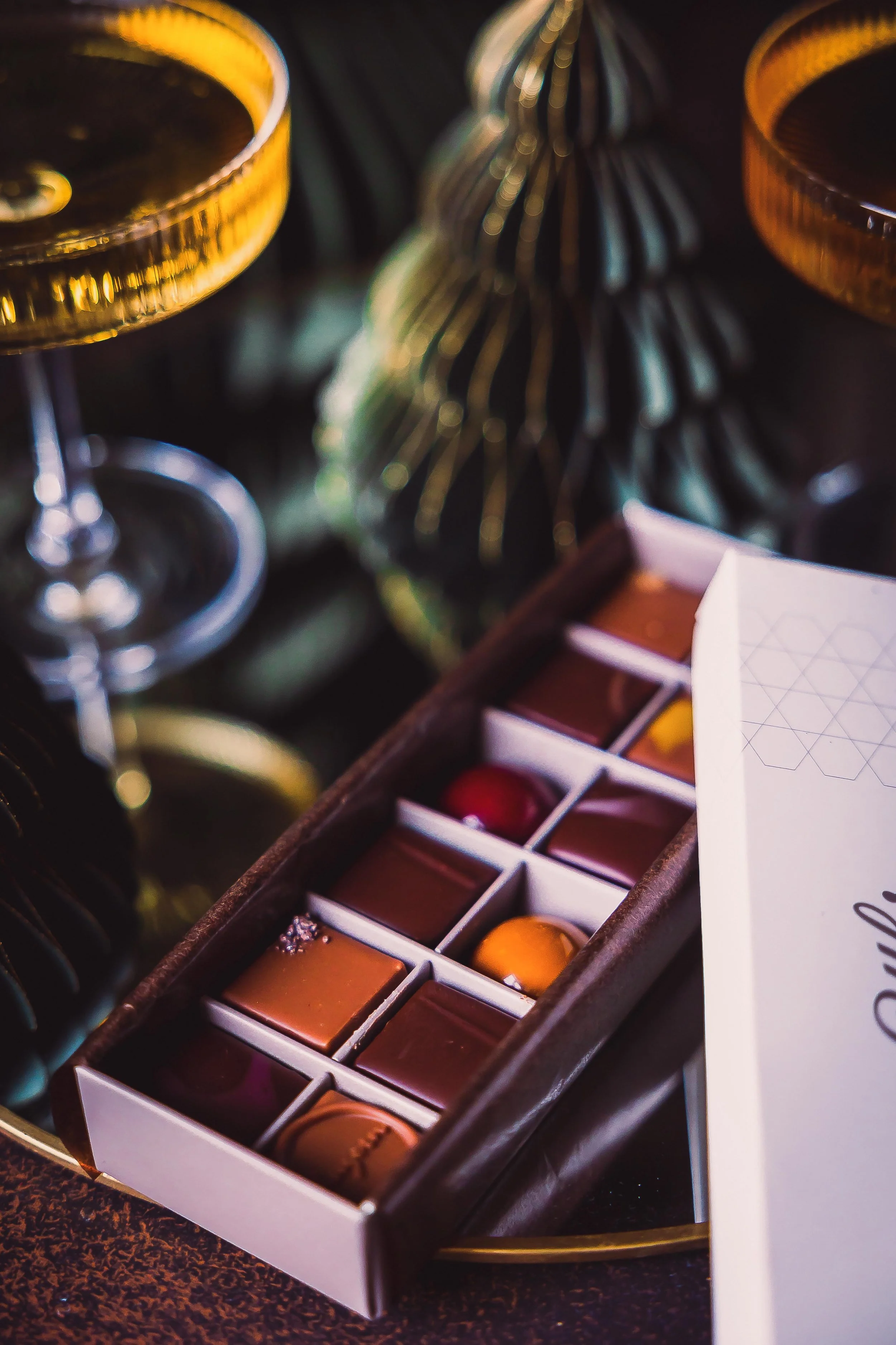 Box of assorted chocolates next to glasses of champagne or wine on a table during a celebration or festive occasion.