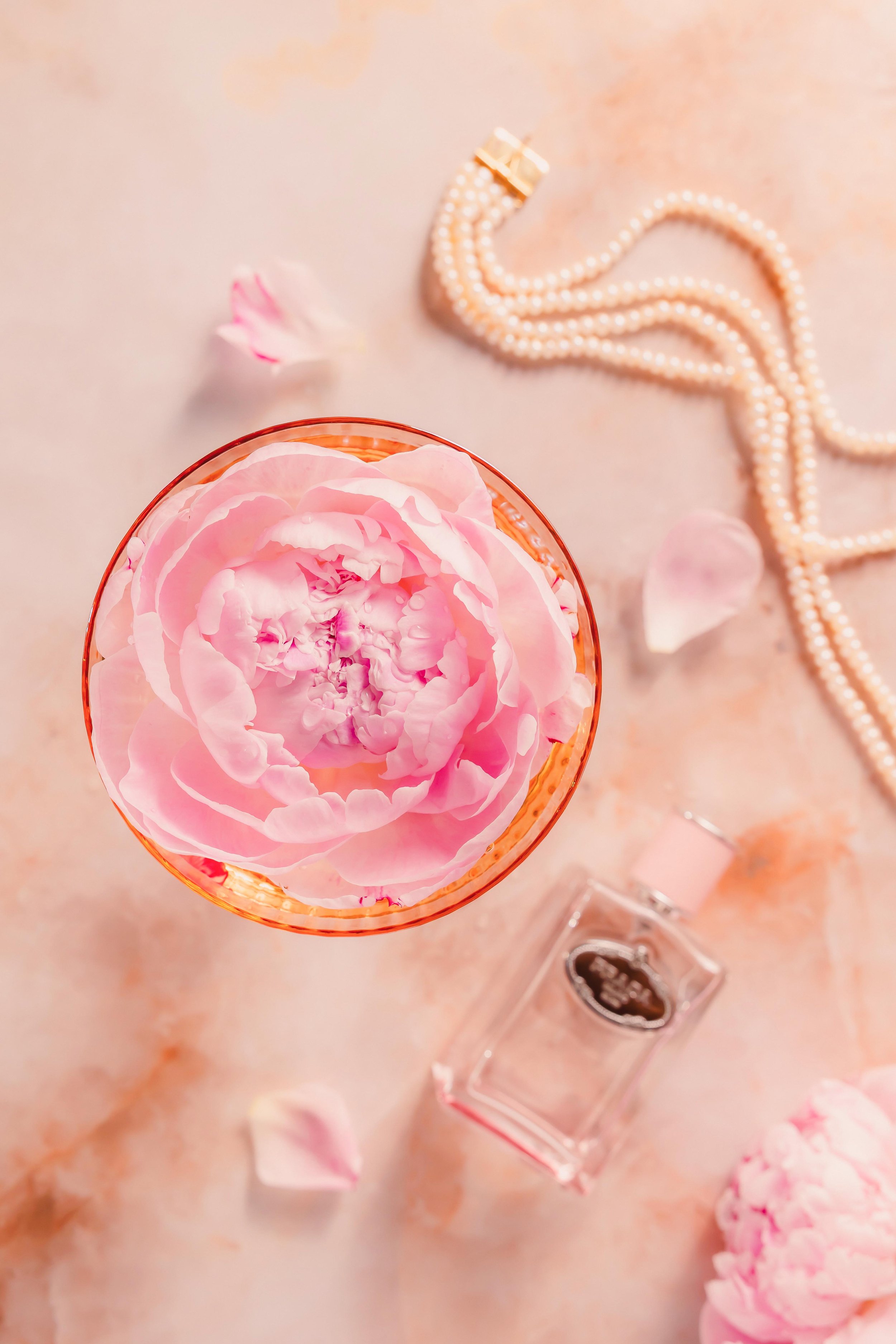 Pink flower in a glass container, surrounded by scattered petals, a pearl necklace, a small perfume bottle, and makeup items on a light-colored surface.