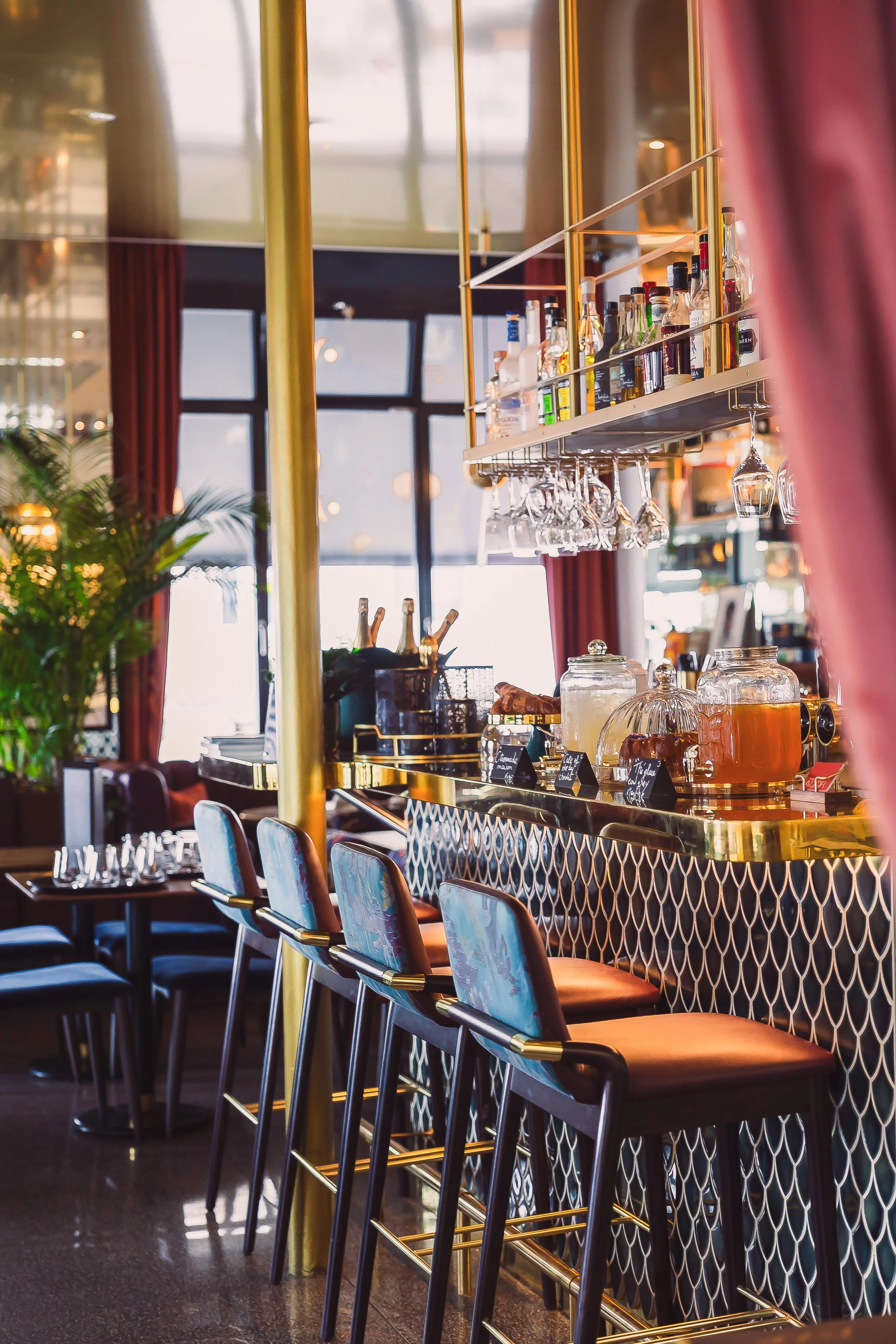 A modern bar with high chairs, a gold-accented bar counter, hanging glasses, and a display of liquor bottles, with large windows and red curtains in the background.