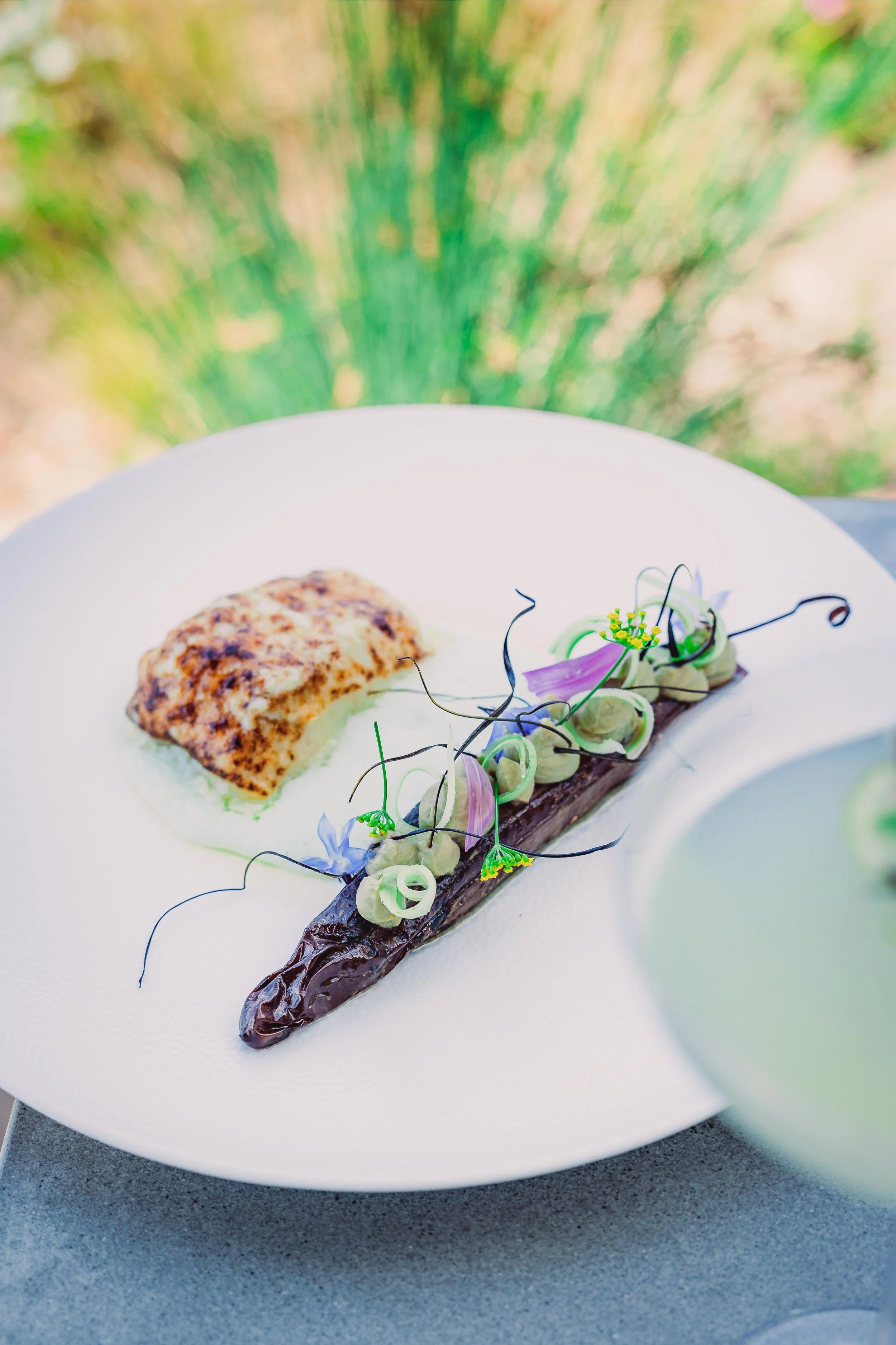A white plate with a piece of grilled fish, a roasted eggplant, and colorful edible flowers and garnishes.