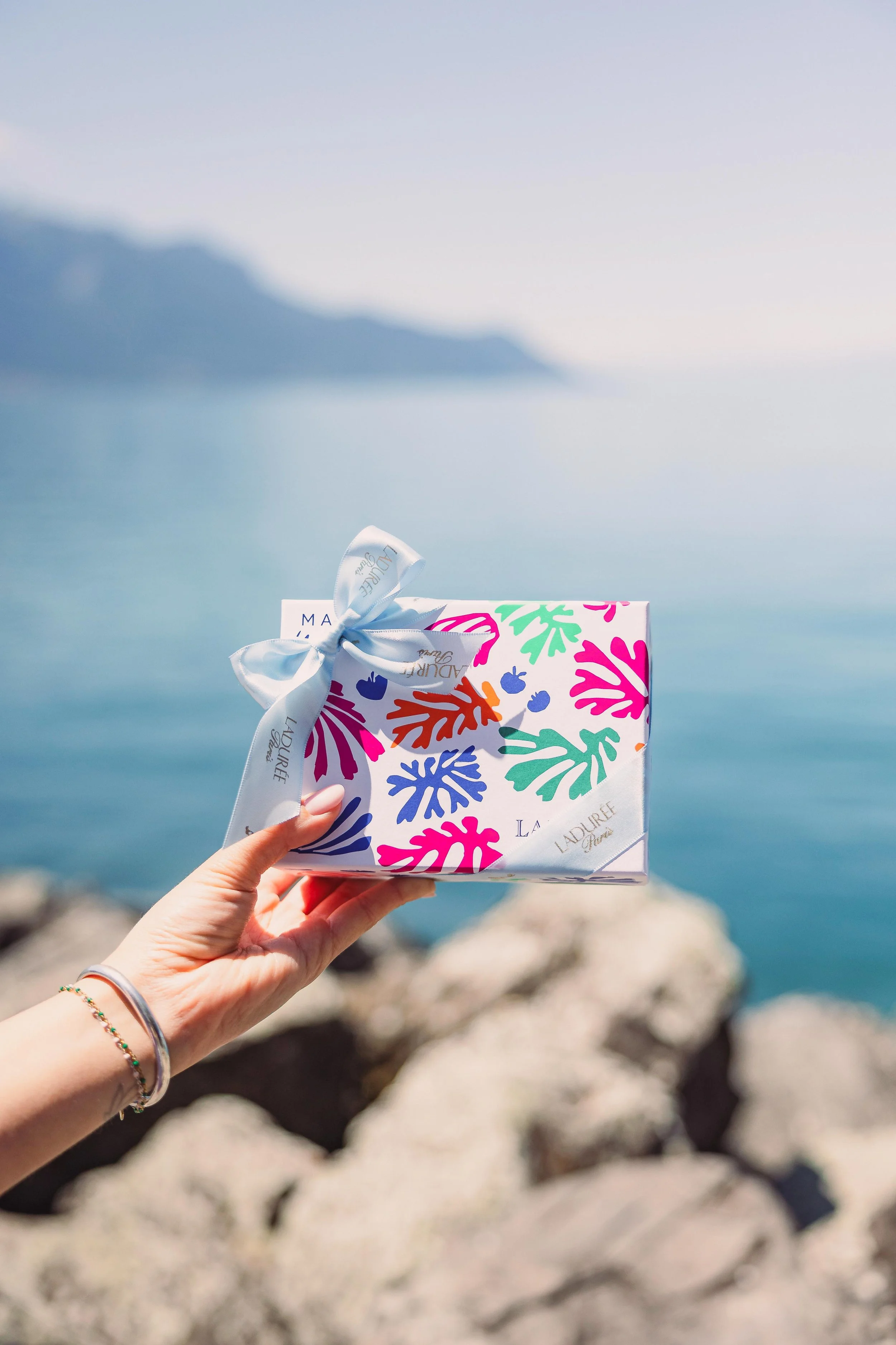 A person holds a colorful gift box with a white ribbon, against a backdrop of a lake and mountains.
