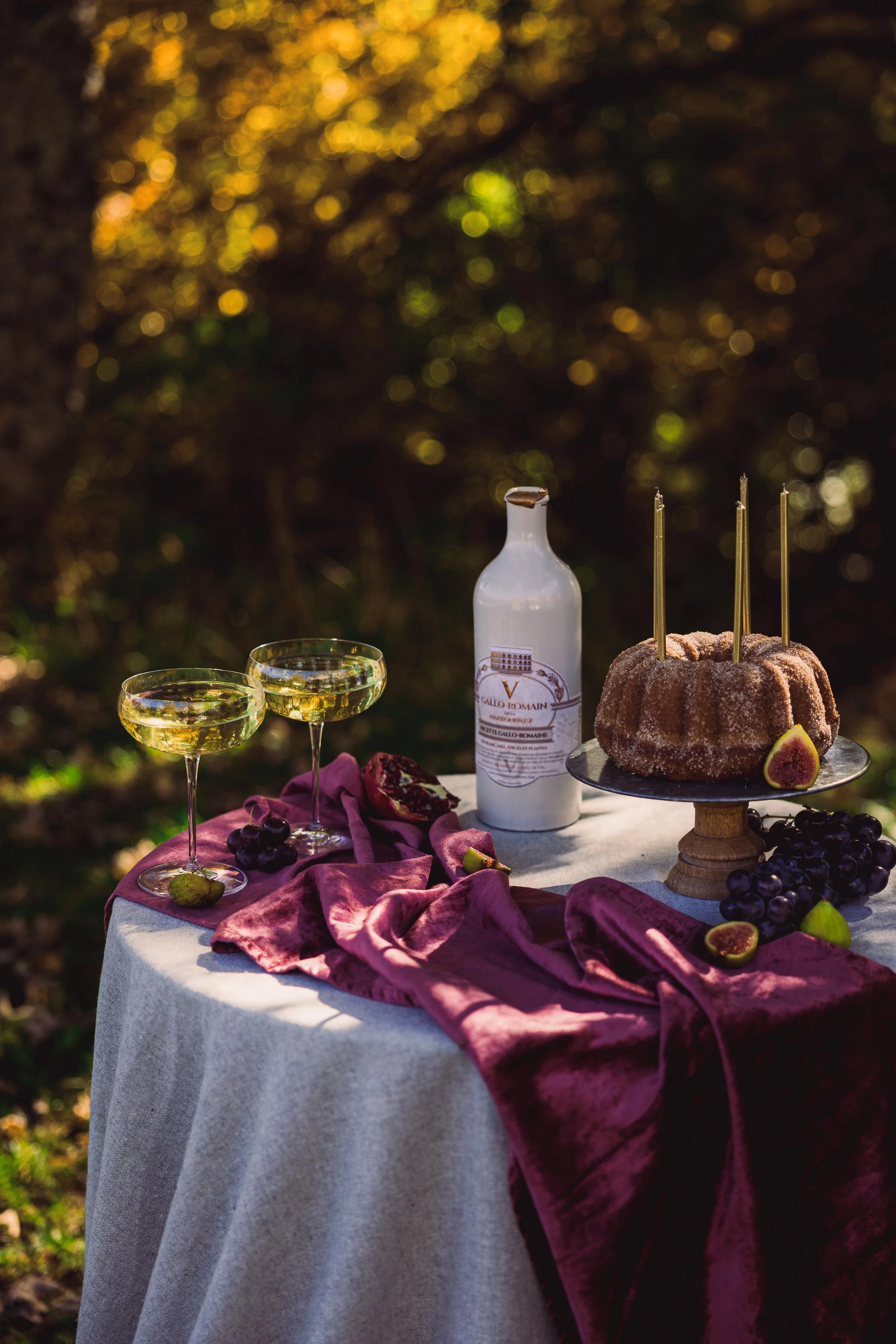 A table with a beige tablecloth and a dark red cloth is set outdoors with two glasses of white wine, a bottle of liquor, a bundt cake with candles, grapes, figs, and a pomegranate on a sunny day.