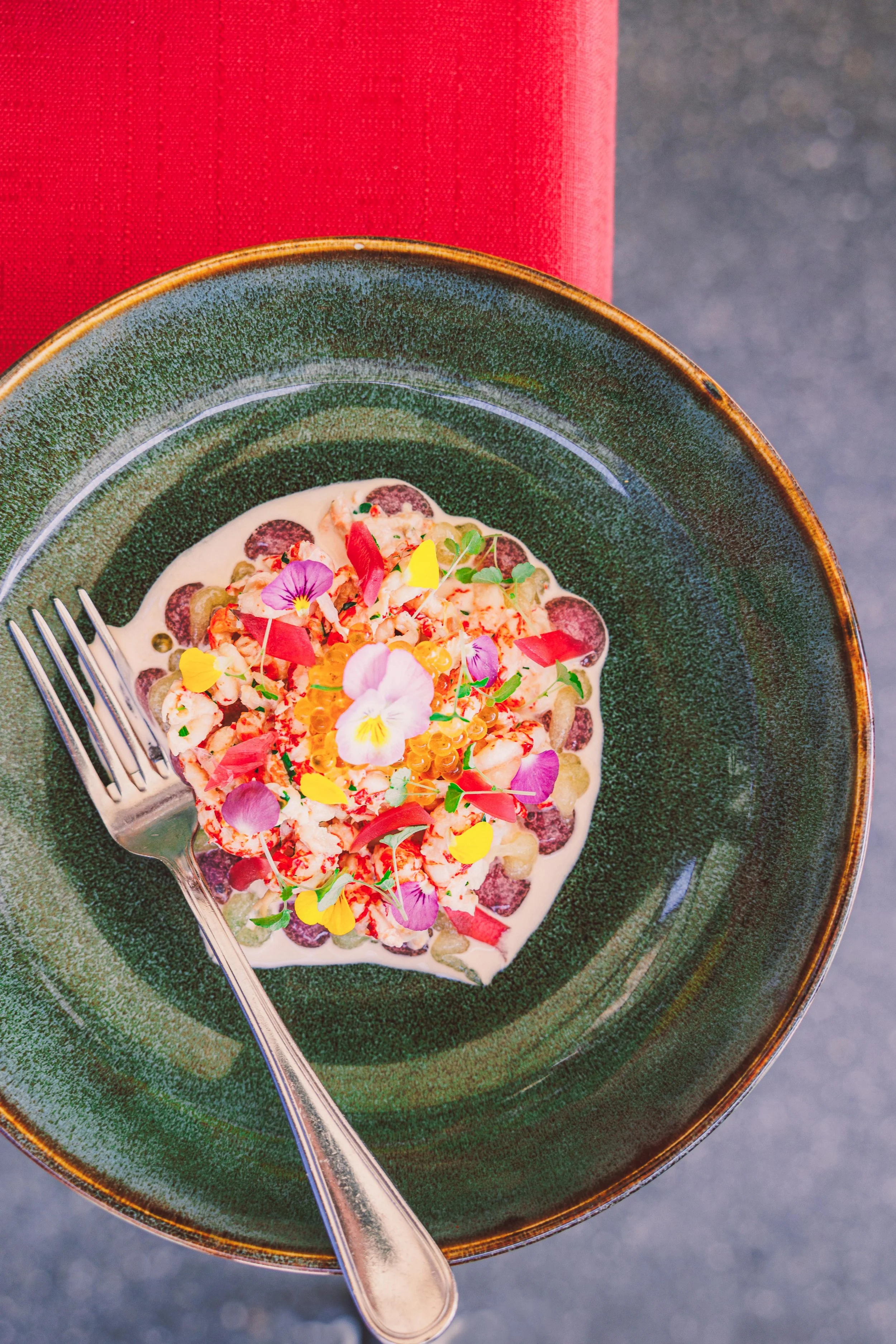 A gourmet dish garnished with edible flowers, served in a green bowl with a fork on the side.