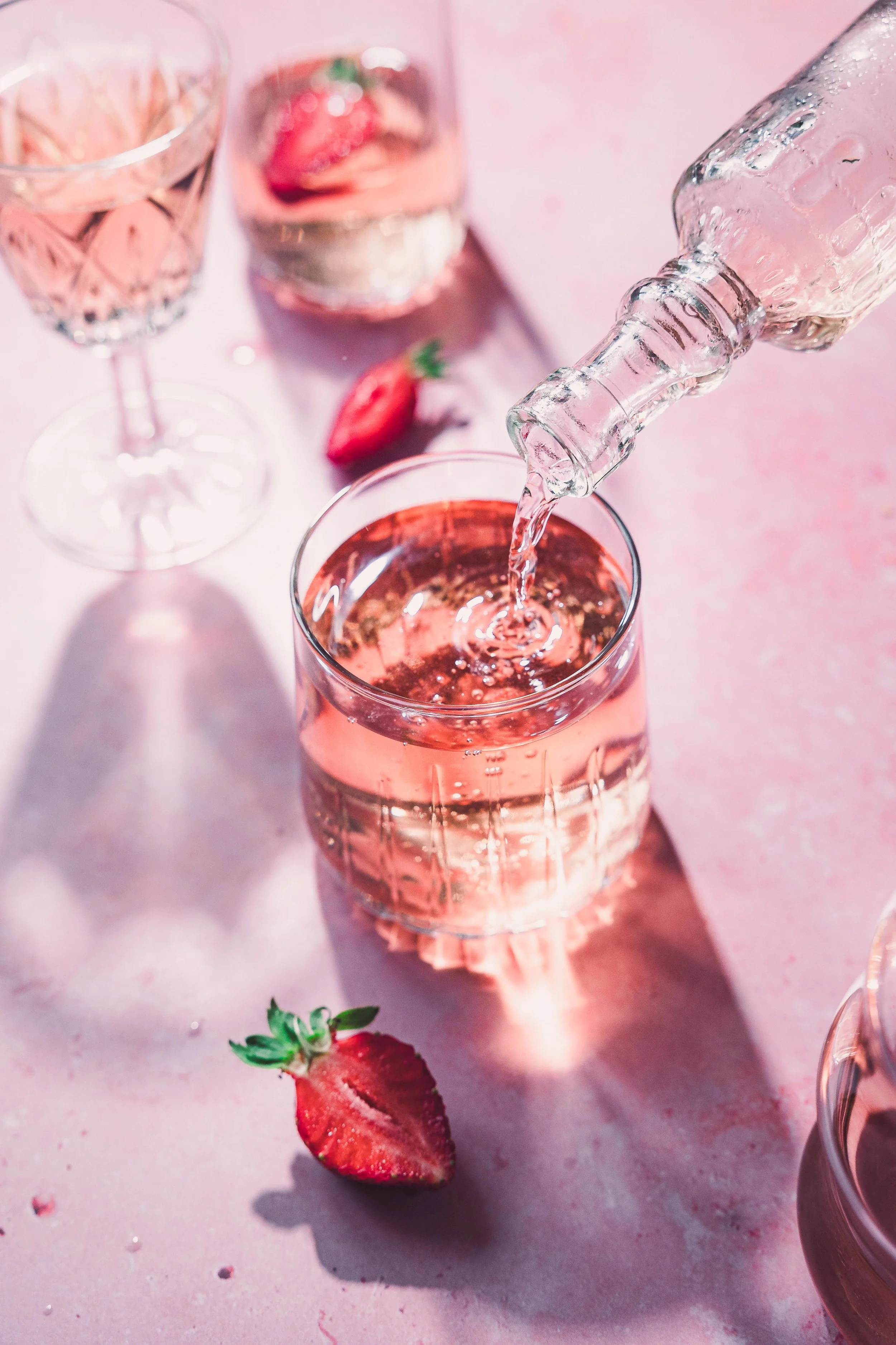 A glass of pink sparkling water being poured from a clear bottle with sliced strawberries on a pink surface.