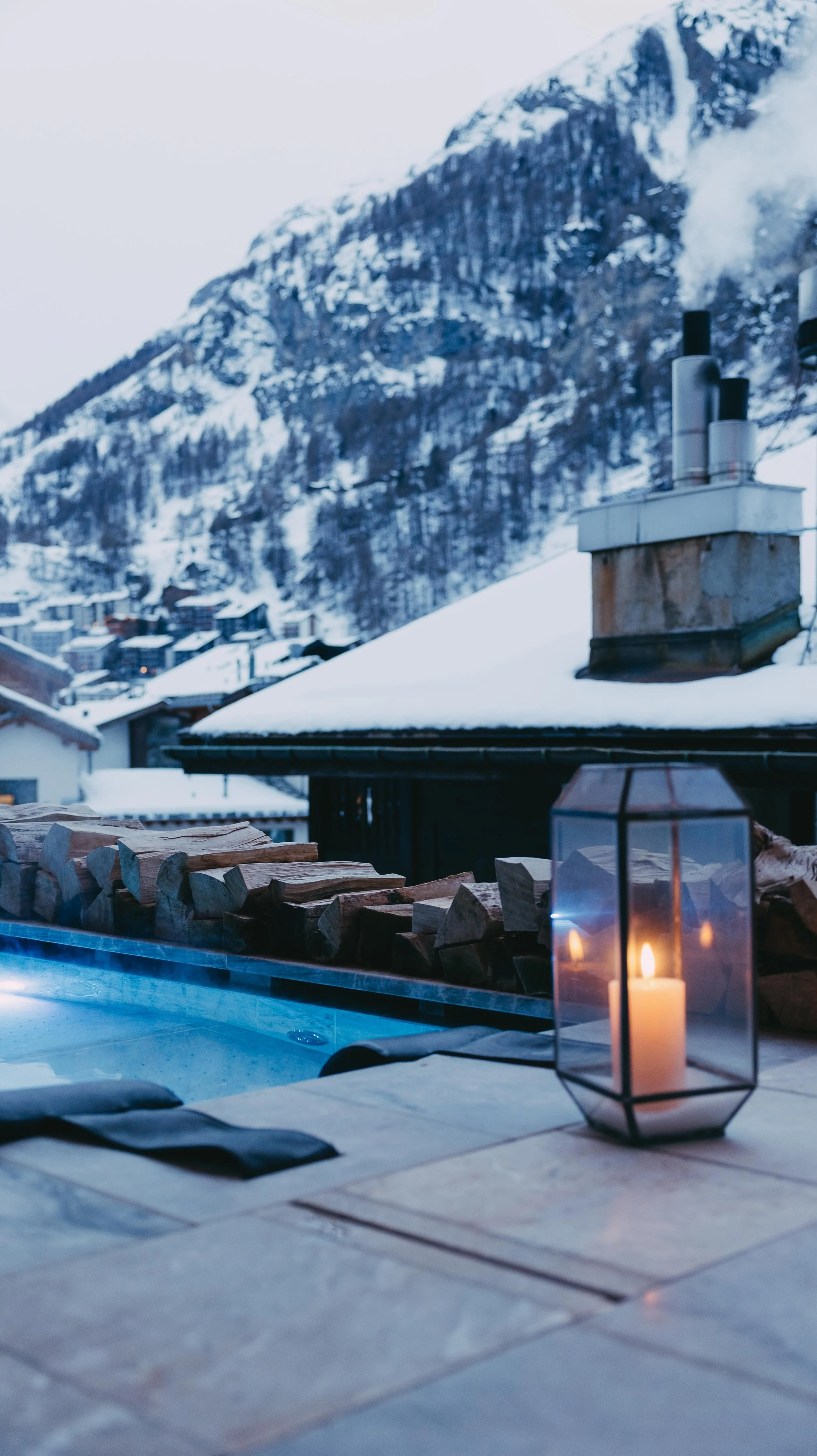 A cozy outdoor hot tub area with candlelight, surrounded by firewood, overlooking snowy mountain and village scenery.