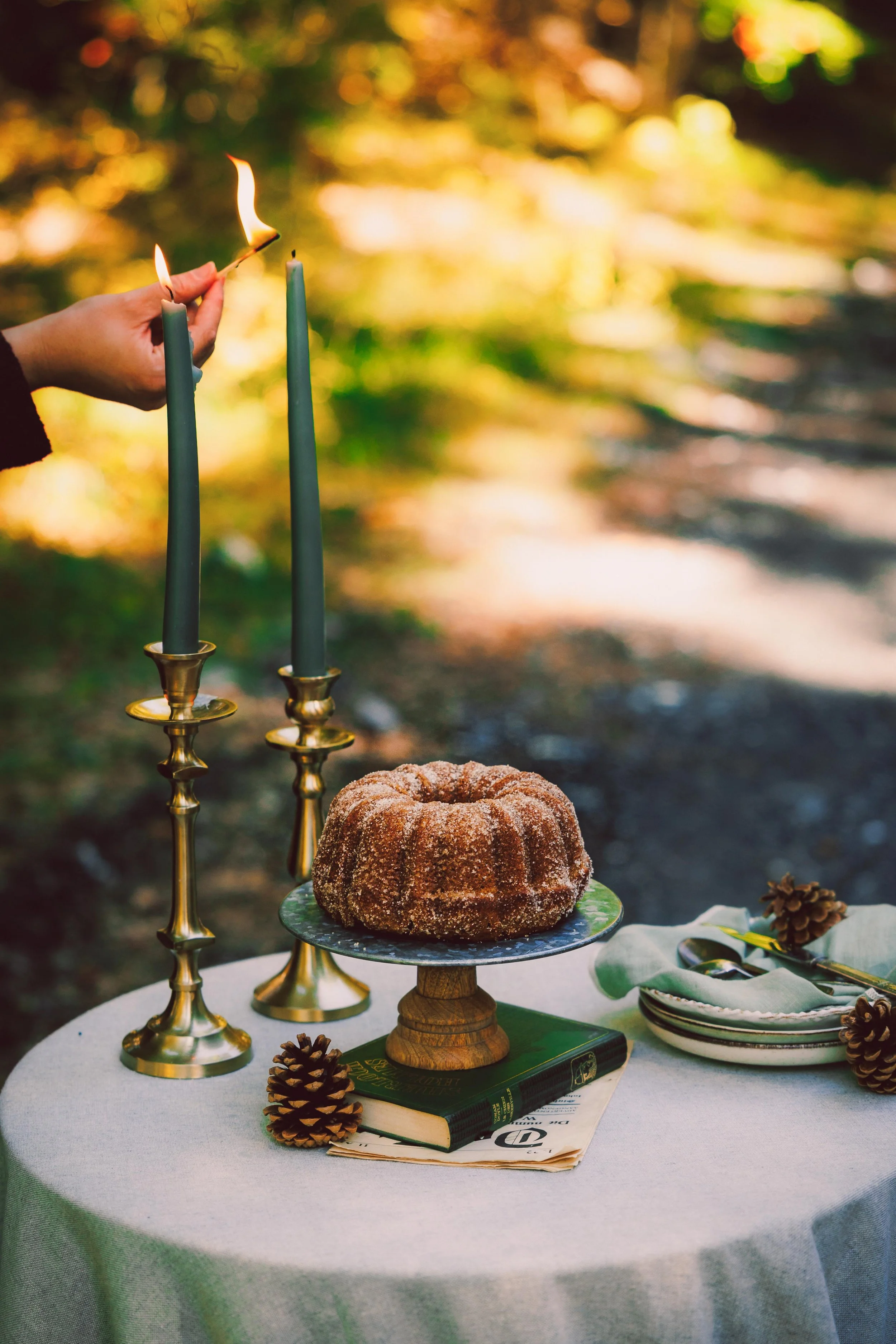 A table set outdoors with a bundt cake on a cake stand, gold candlesticks with green candles, pinecones, a book, and a plate with utensils, with a background of trees and greenery.