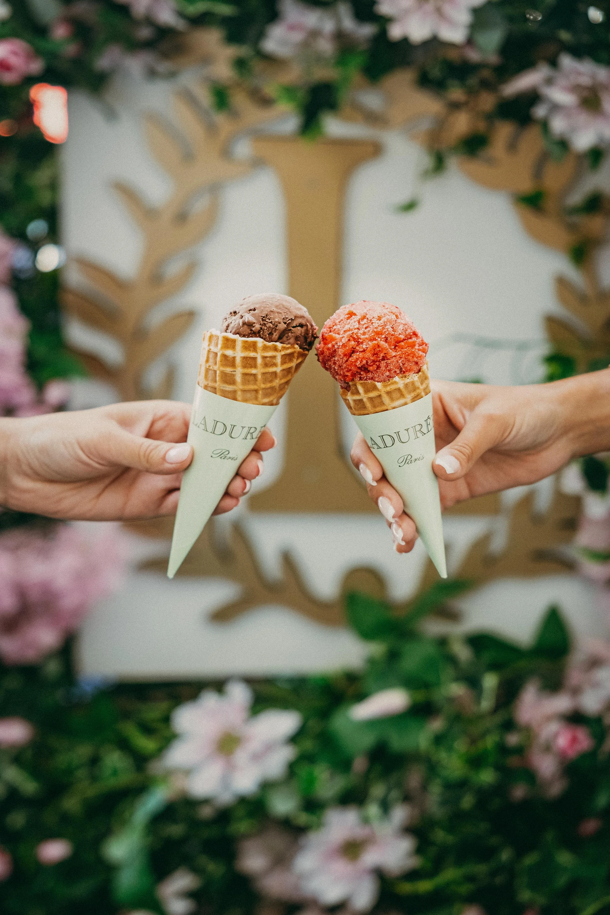 Two people holding ice cream cones with the word "LADUREE" on the wrappers, with a decorative background featuring a gold-colored tree and pink flowers.
