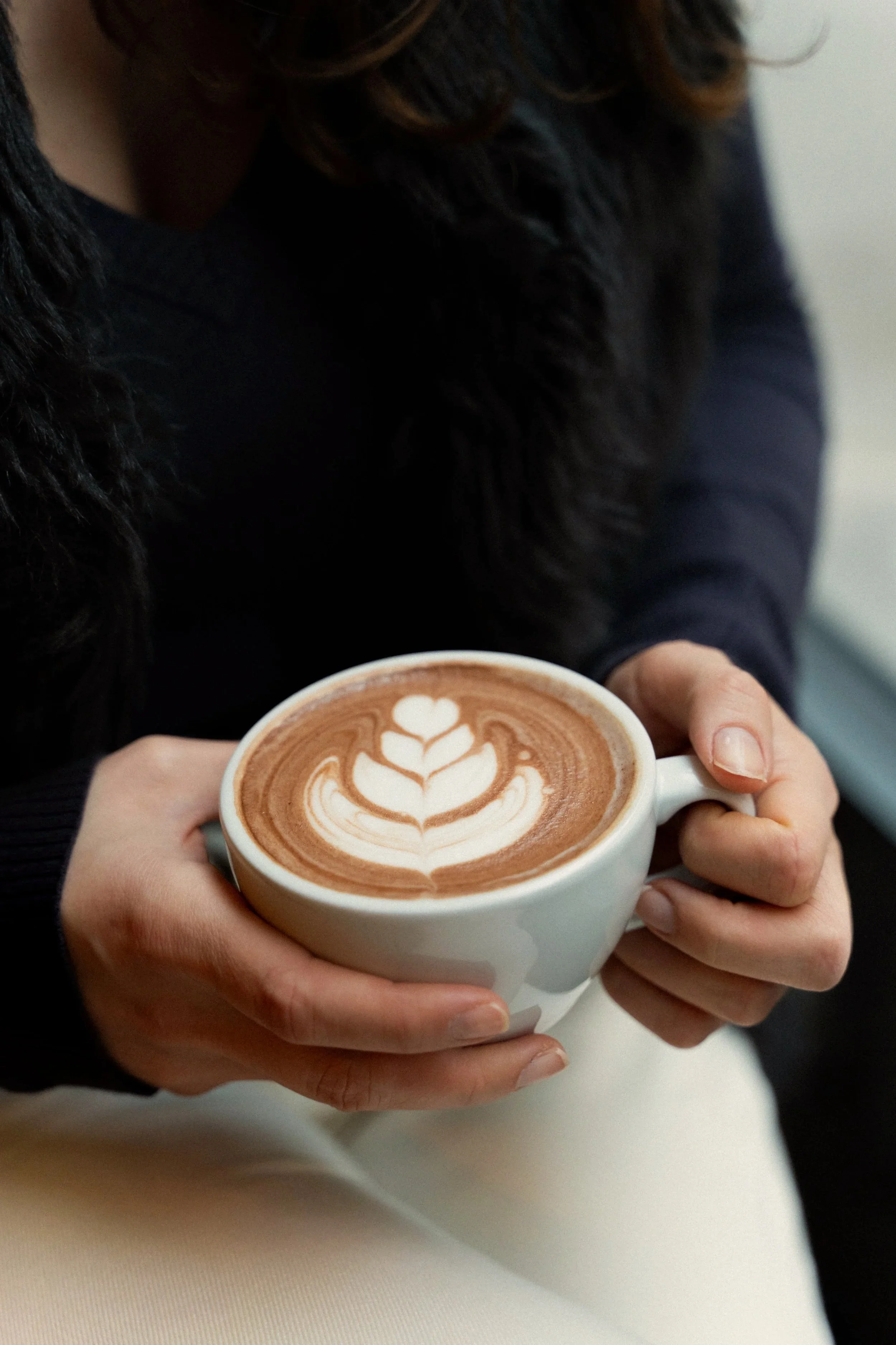 Person holding a cup of coffee with latte art in the shape of a heart. The person is wearing a dark sweater with fur detail and has long hair.