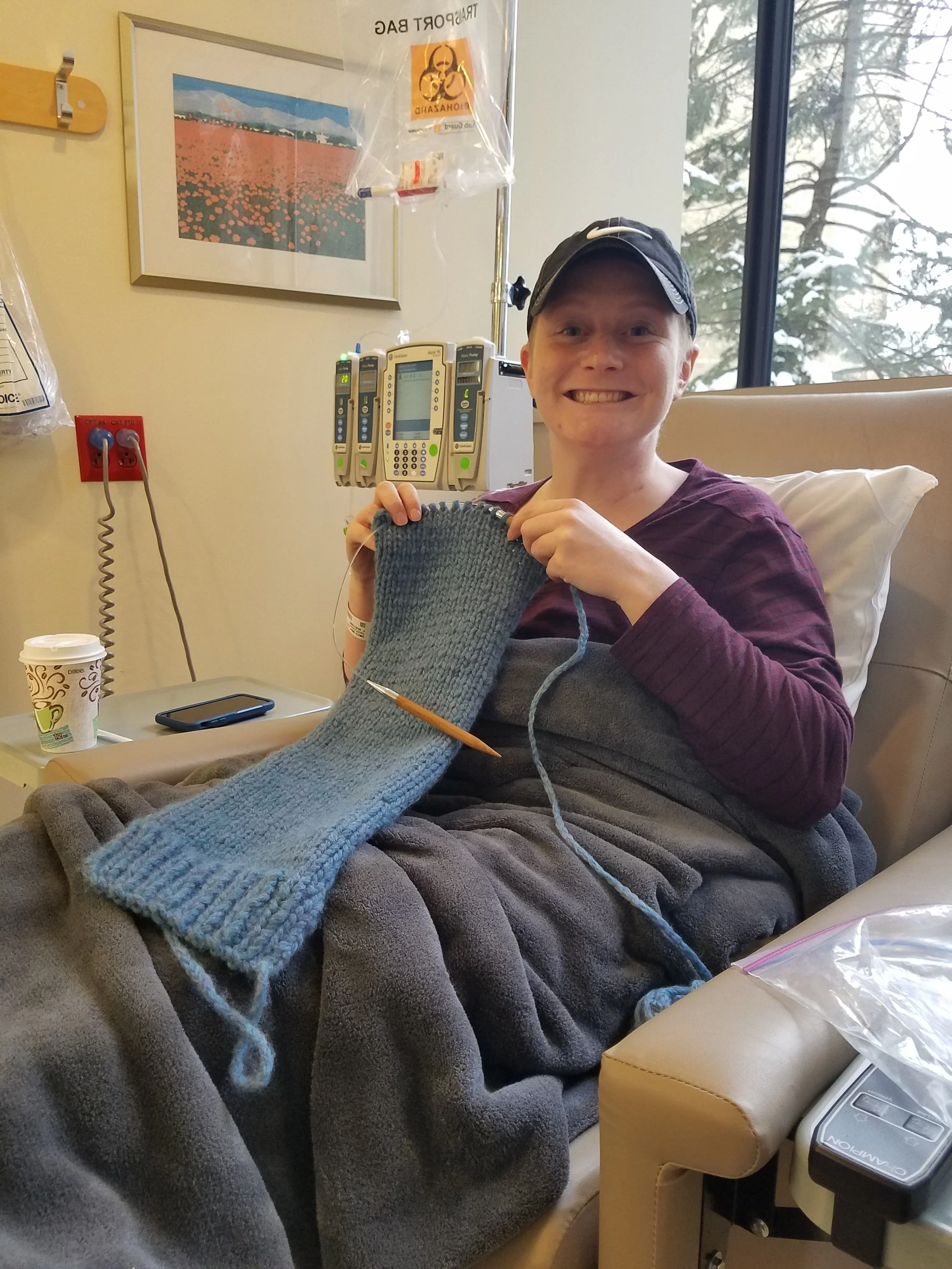 Person receiving chemotherapy and knitting with a gray yarn, smiling at the camera. Items on a side table include a coffee cup and a smartphone. Medical equipment and a window looking out onto snow-covered trees are visible in the background.