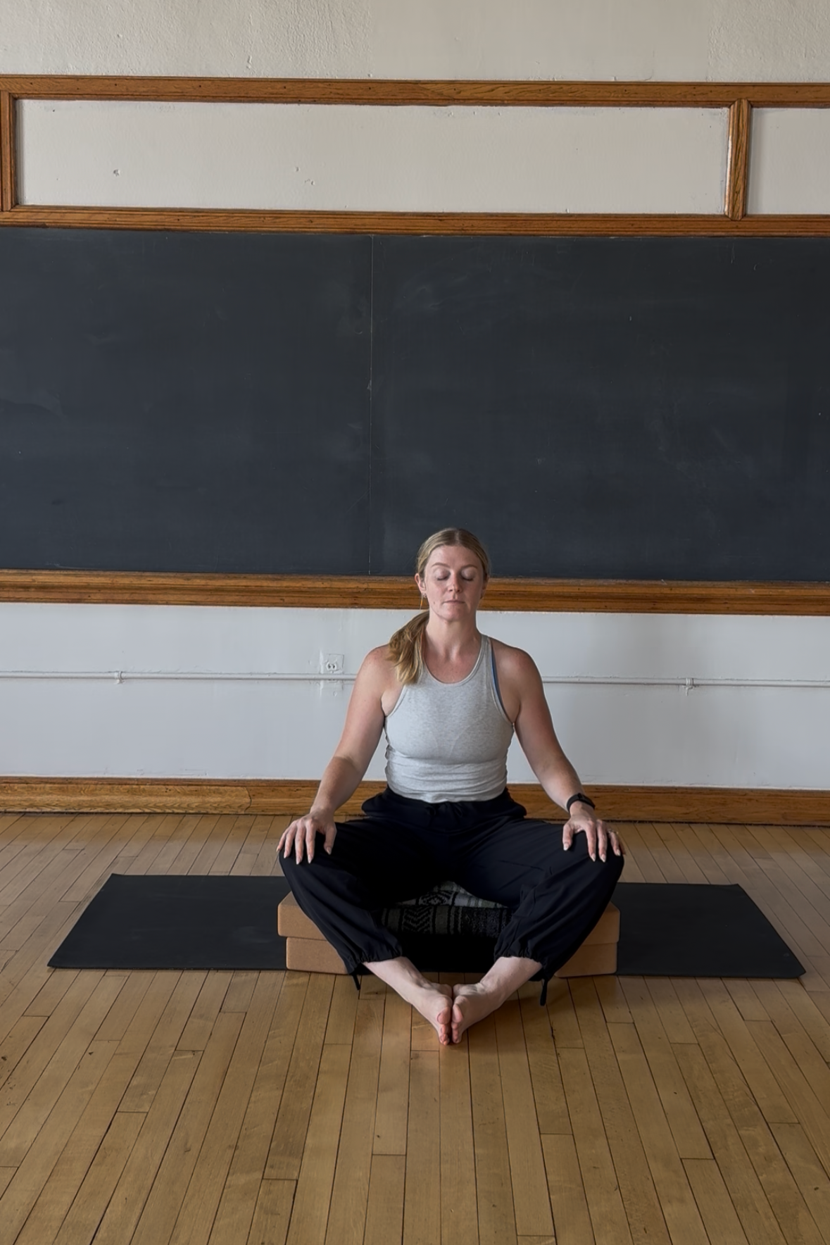 A woman practicing yoga in a seated meditation pose on a yoga mat in a classroom or studio with a black chalkboard and wooden floor.