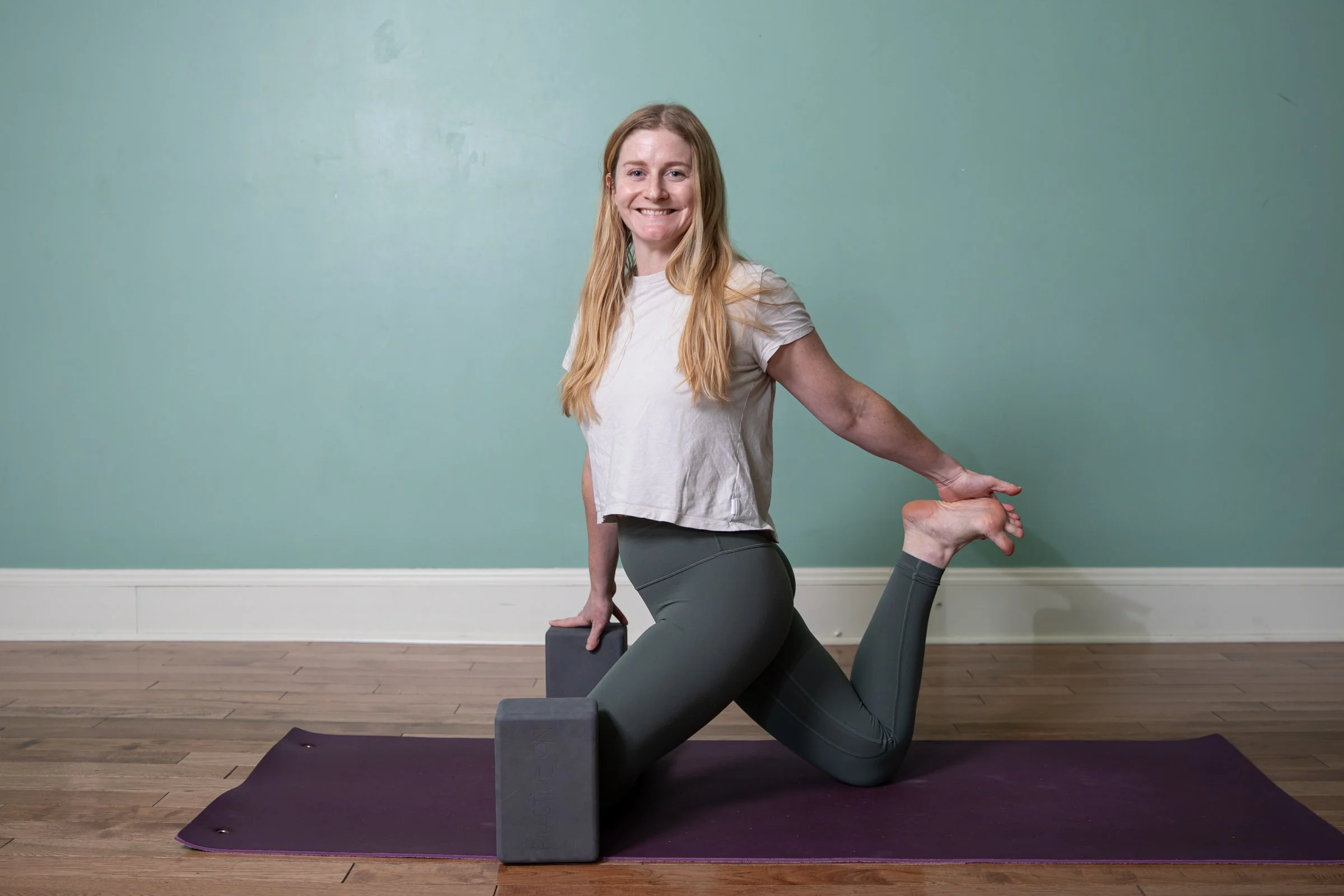 A woman with long blonde hair smiling in a yoga pose, balancing on her right knee with her left leg extended back, holding her left foot with her left hand, and resting her right hand on a yoga block, in front of a blue-green wall with a white baseboard.