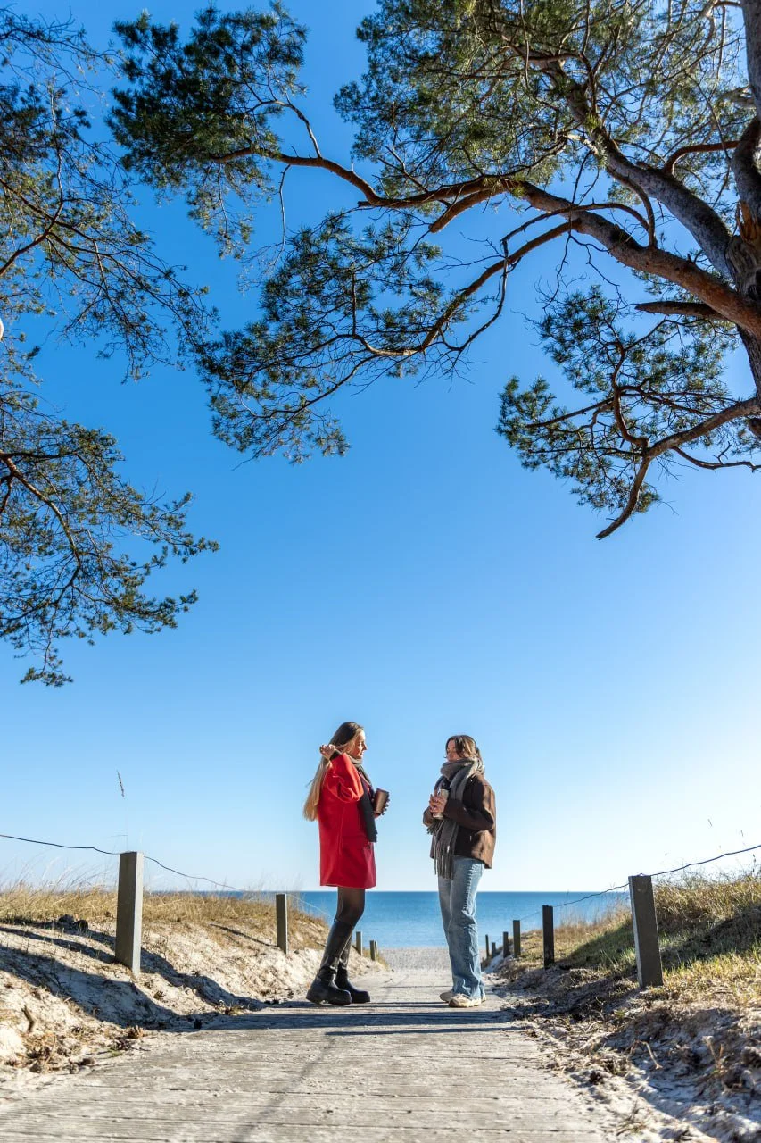 Two women talking on a pathway near the beach, under large trees with clear blue sky overhead.