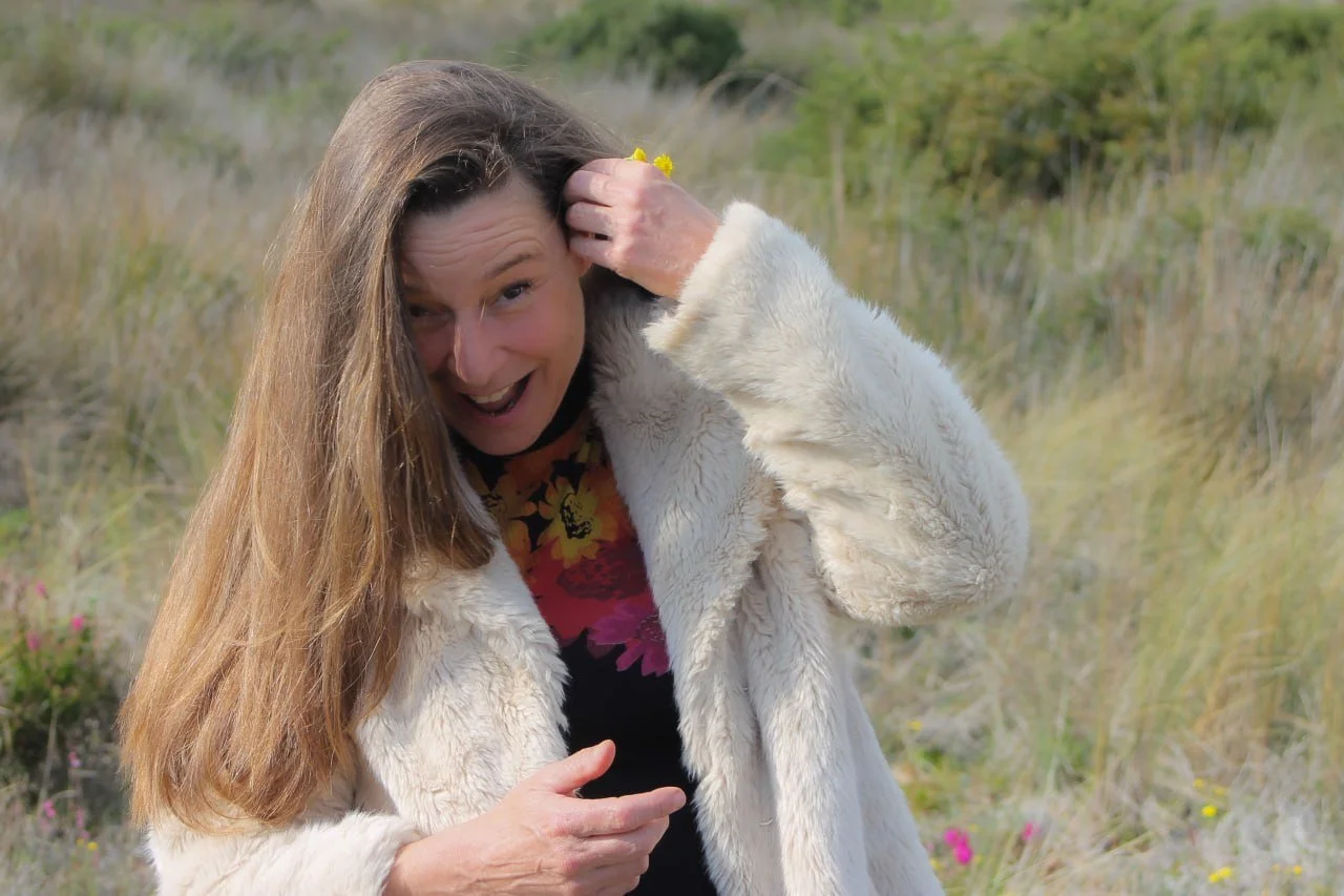 A woman with long brown hair wearing a white faux fur coat and a colorful floral top, smiling outdoors in a grassy field with wildflowers.