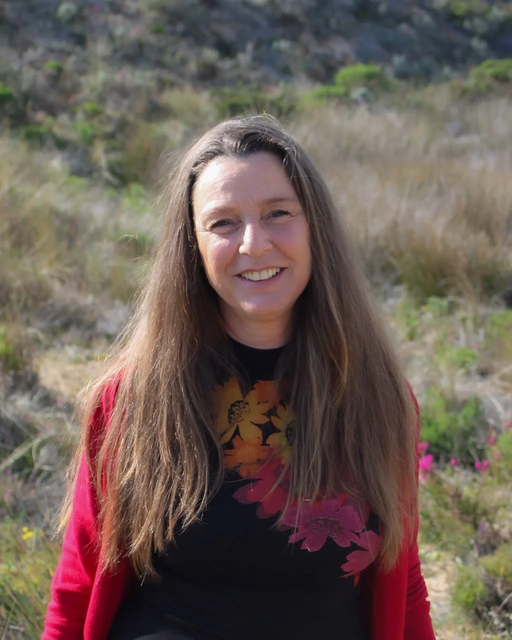 A woman with long brown hair smiling outdoors in a natural setting with green bushes and rocky hills in the background.