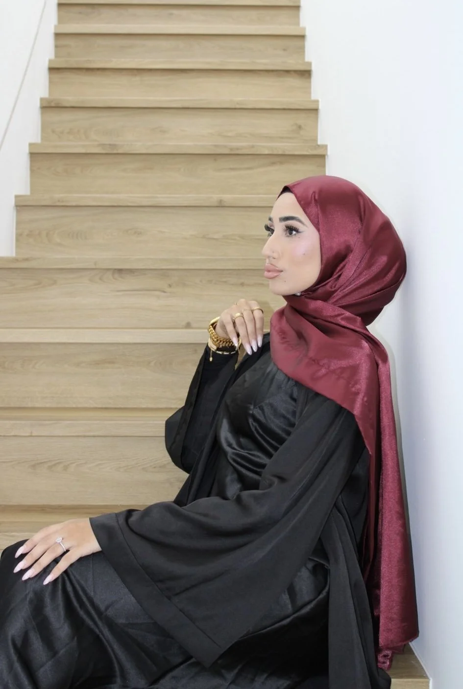A woman in graduation robes and a maroon hijab sitting on a step, looking to the side with jewelry on her hand.