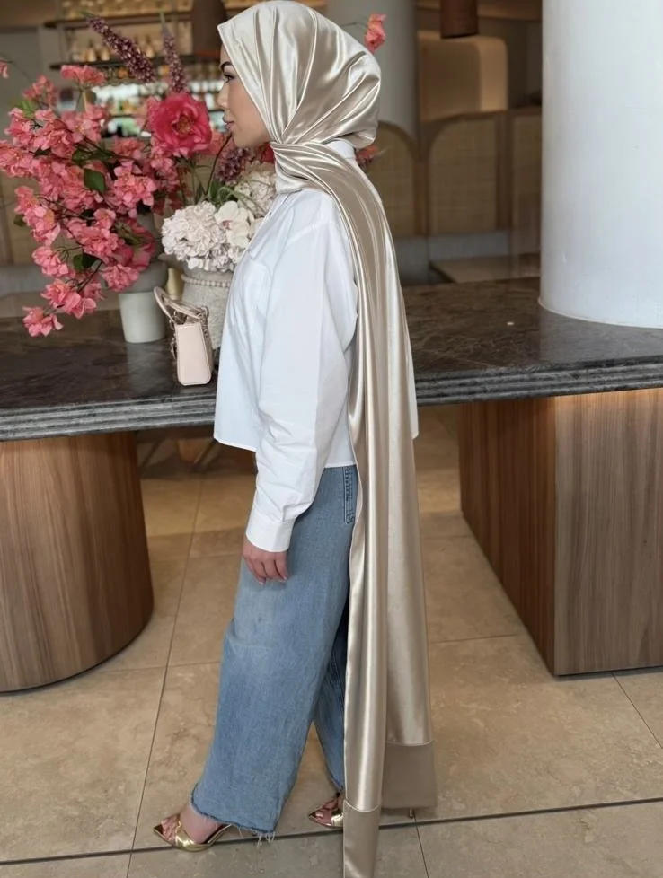 A woman wearing a shiny beige hijab, a white shirt, and light blue jeans standing next to a marble-topped wooden counter with a large bouquet of pink and white flowers on it in a modern interior setting.