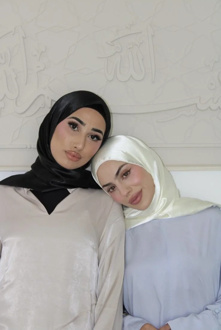 Two women wearing hijabs, one in black and the other in cream-colored satin, posing closely together against a wall with Islamic calligraphy.