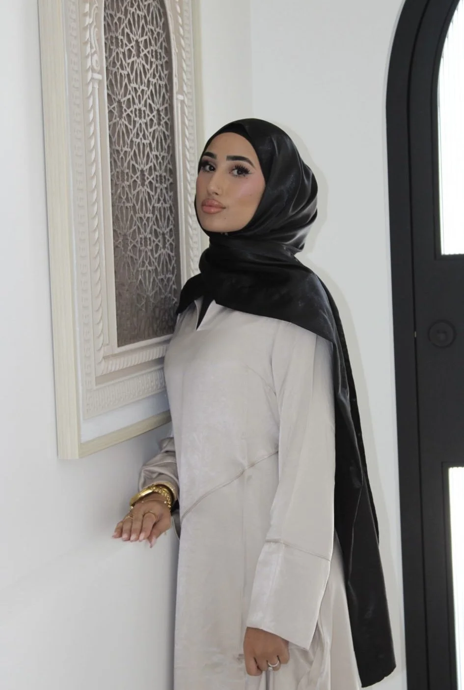 A woman wearing a black headscarf, a beige dress, and gold jewelry, standing by a decorative wall piece and a black door in a well-lit interior.