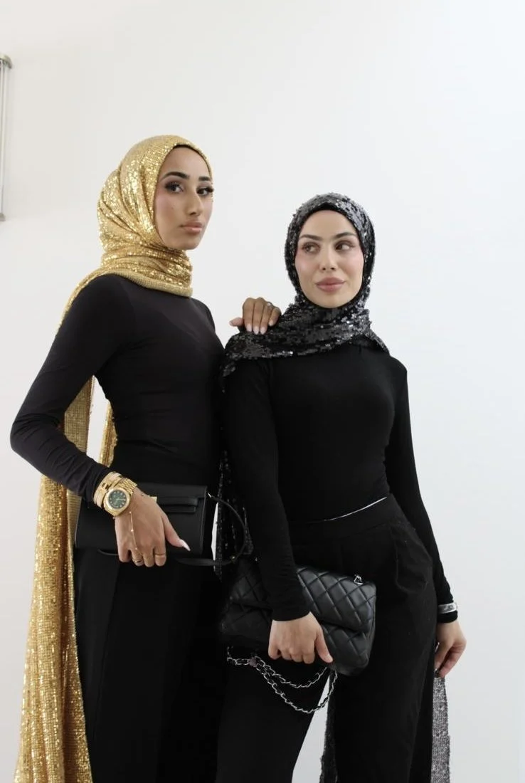 Two women wearing black outfits and sequin headscarves, one in gold and the other in silver, standing against a plain white background.
