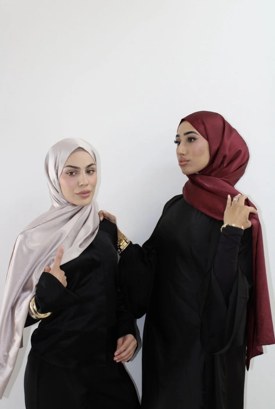 Two women wearing hijabs and black outfits, standing against a white background.