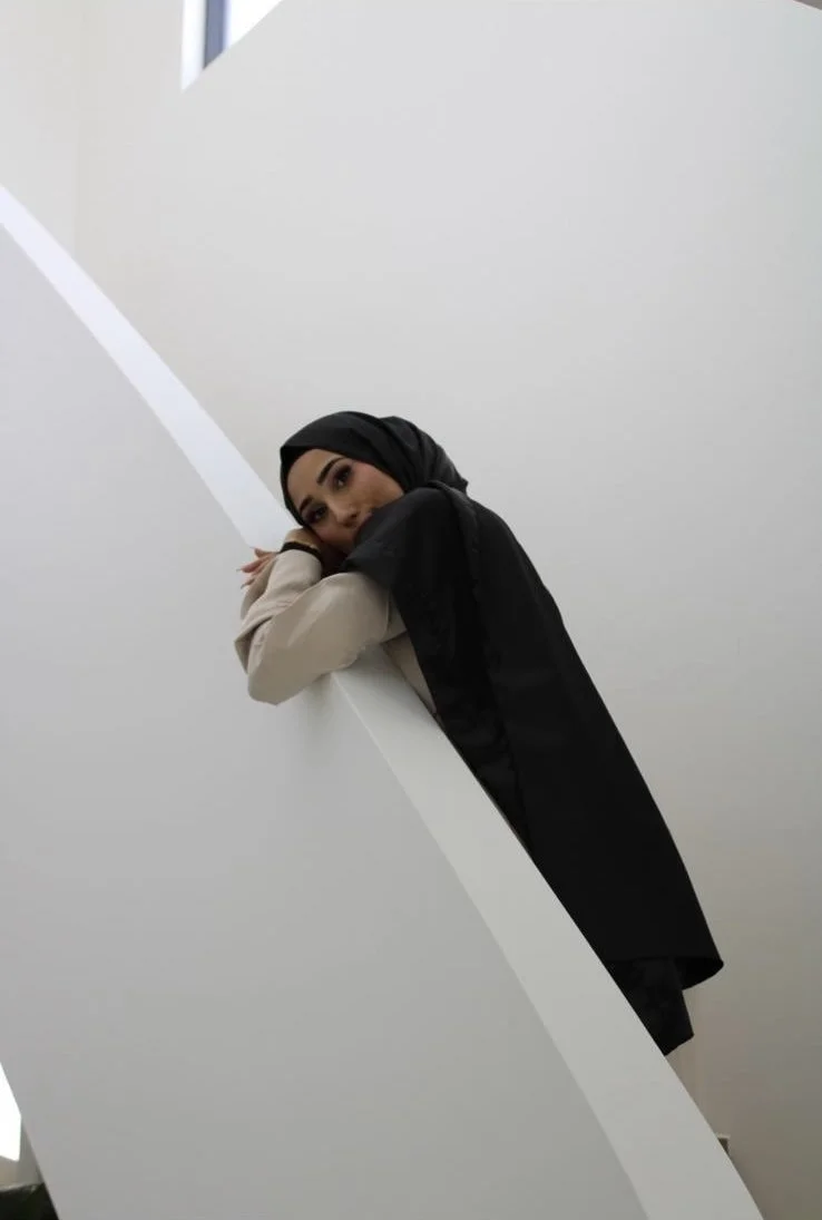 A young woman with dark eyebrows and makeup, wearing a black hijab, leaning on a white curved wall with her head resting on her crossed arms, in a modern indoor setting.