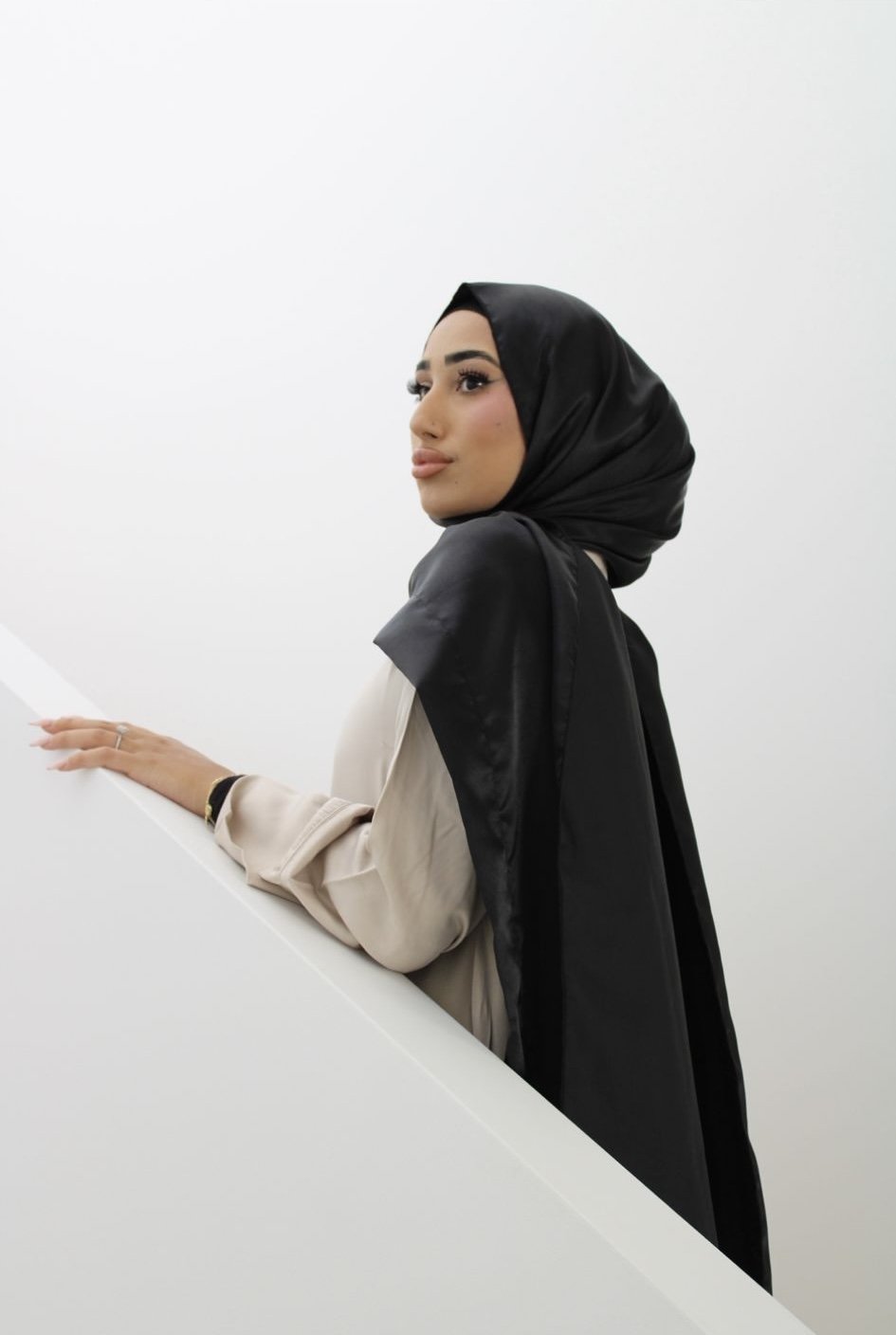 A woman wearing a black hijab and beige blouse, standing against a white wall with her hand resting on a railing, looking to the side.