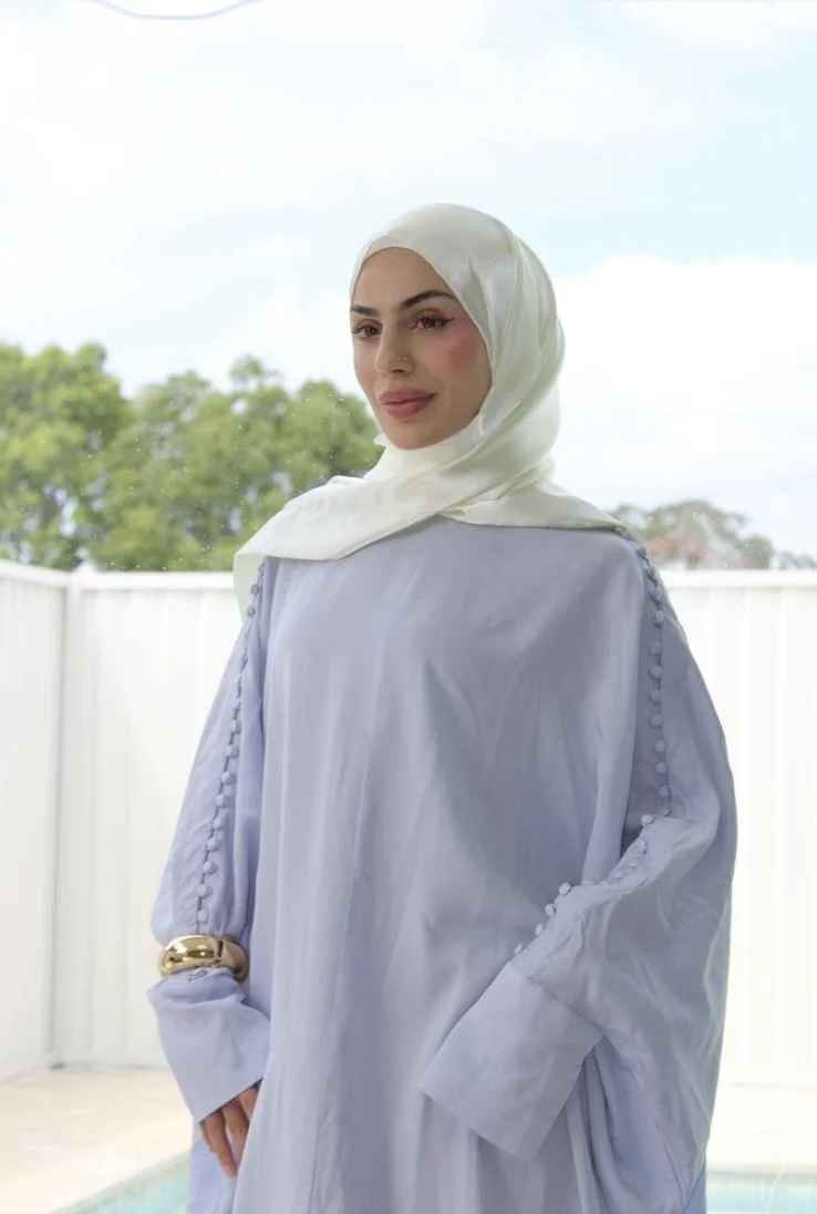 A woman wearing a white hijab and a light blue dress with decorative buttons on the sleeves, standing outdoors with a white fence and green trees in the background.