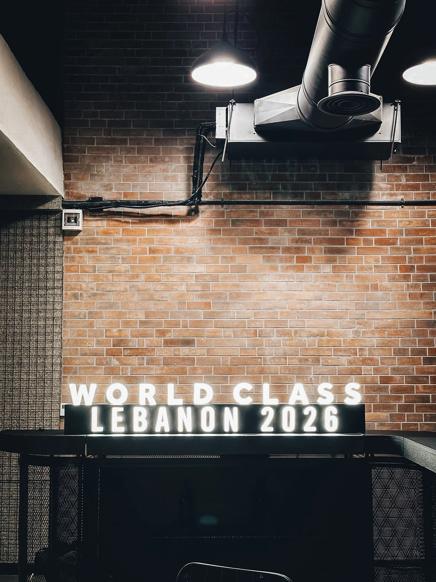 Bright illuminated sign on a black surface reads: 'WORLD CLASS LEBANON 2026' against a brick wall with industrial lighting and ventilation duct overhead.