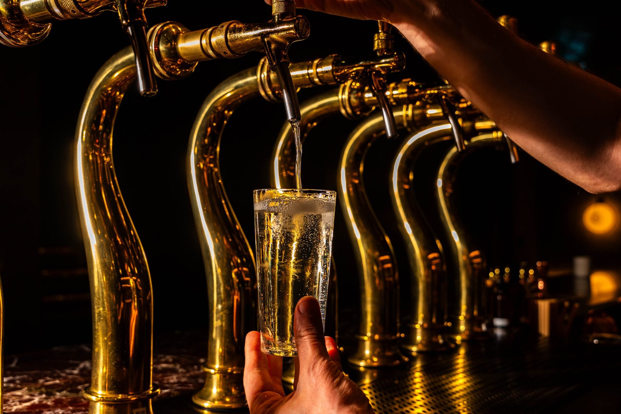 A hand holding a glass of beer under a gold beer tap in a bar or brewery setting.