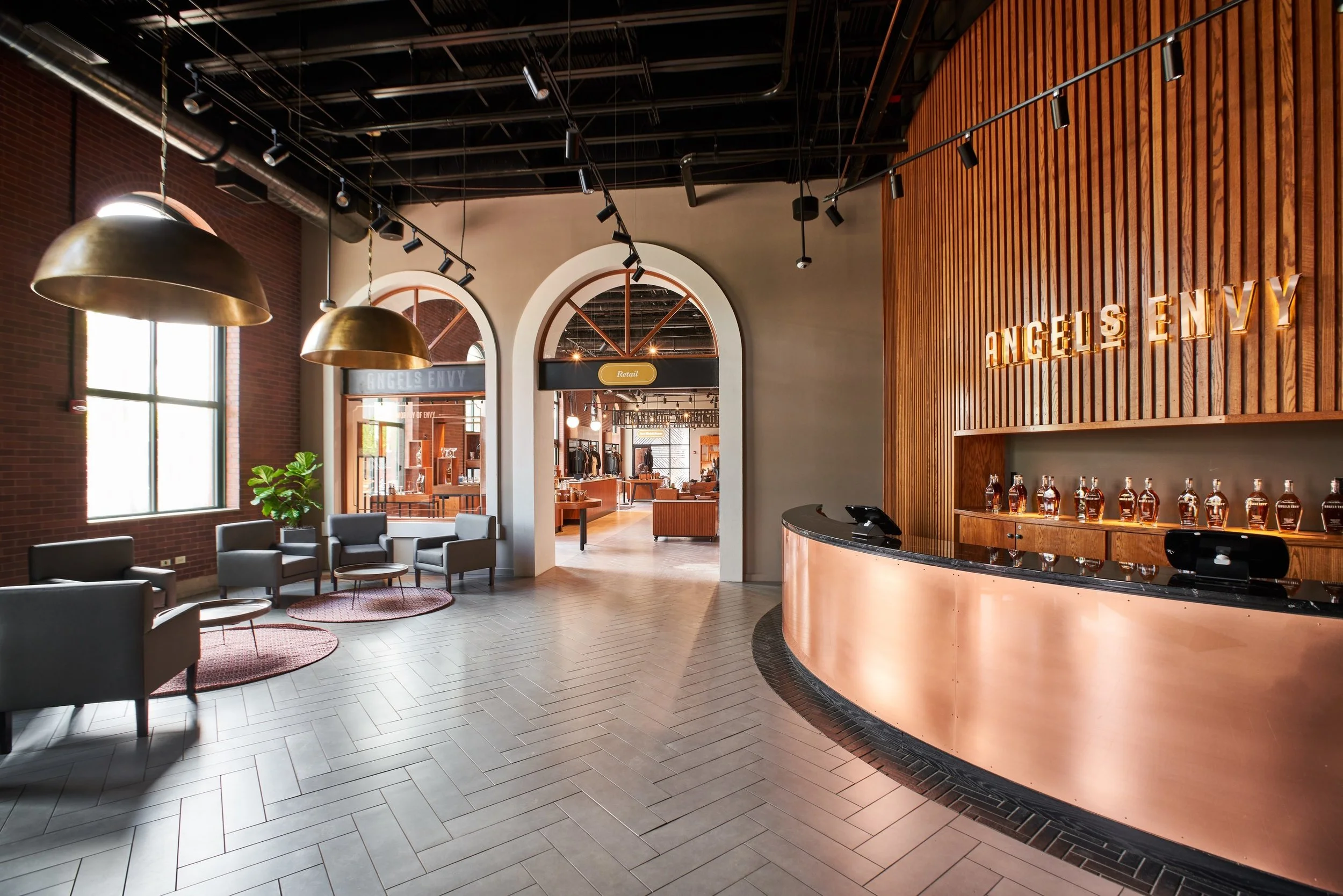 Modern hotel lobby with seating area, large windows, brass pendant lights, wood accents, and a reception desk with bottles of alcohol displayed.