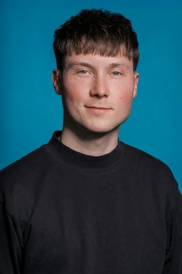 A young, white, male-presenting person is facing the camera with a subtle smile. They are against a blue background, with a black high necked top on. Their hair is short, dark brown. They are wearing hearing aids in both ears.