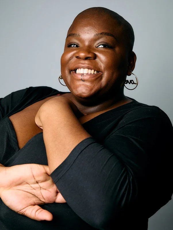A smiling dark-skinned, female-presenting person with hoop earrings, bald, with a black long sleeved top. The person is smiling widely and touching their chest with their left hand, with their right hand moving across their body expressively.