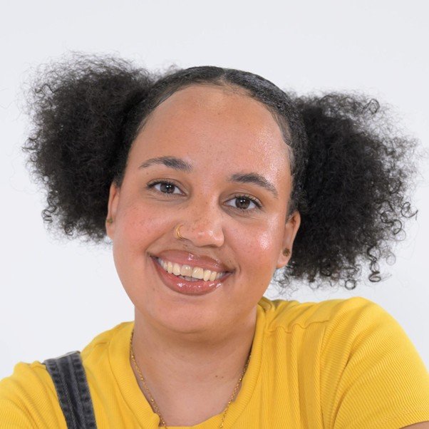 A young brown-skinned, female presenting person is smiling in a friendly manner.  Their curly hair is in two bunches on either side of their head. They are wearing a bright yellow top and looking directly at the camera.