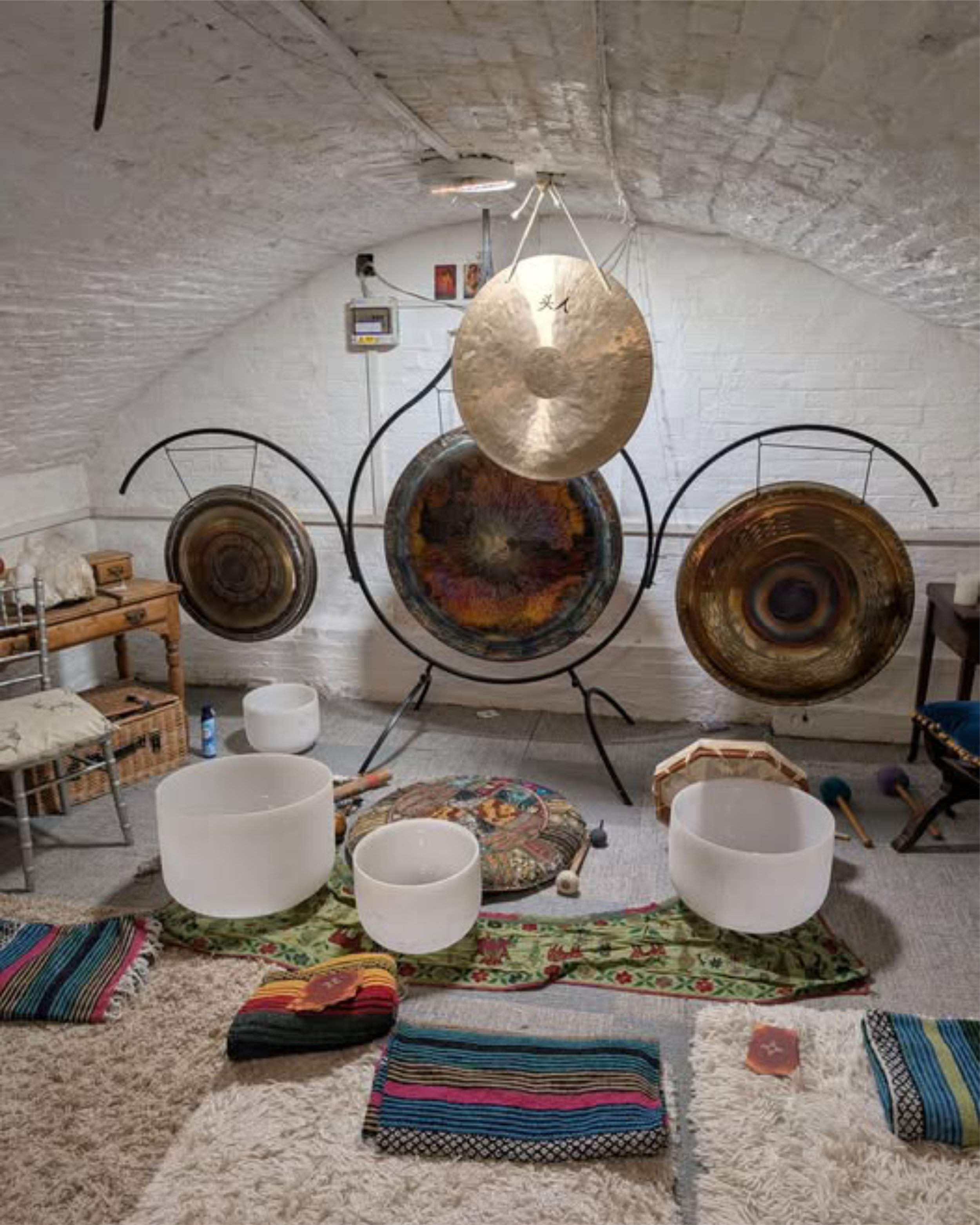 A room with a musical setup including large gongs, white singing bowls, and various cushions, rugs, and colorful textiles on a fluffy white carpet.