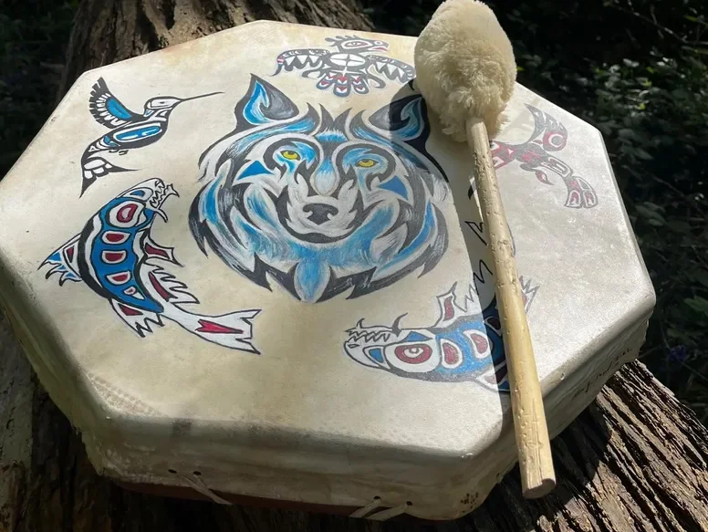 Octagonal box with tribal-style wolf and fish artwork, drumstick with fluffy top resting on it, outdoors on a tree trunk.